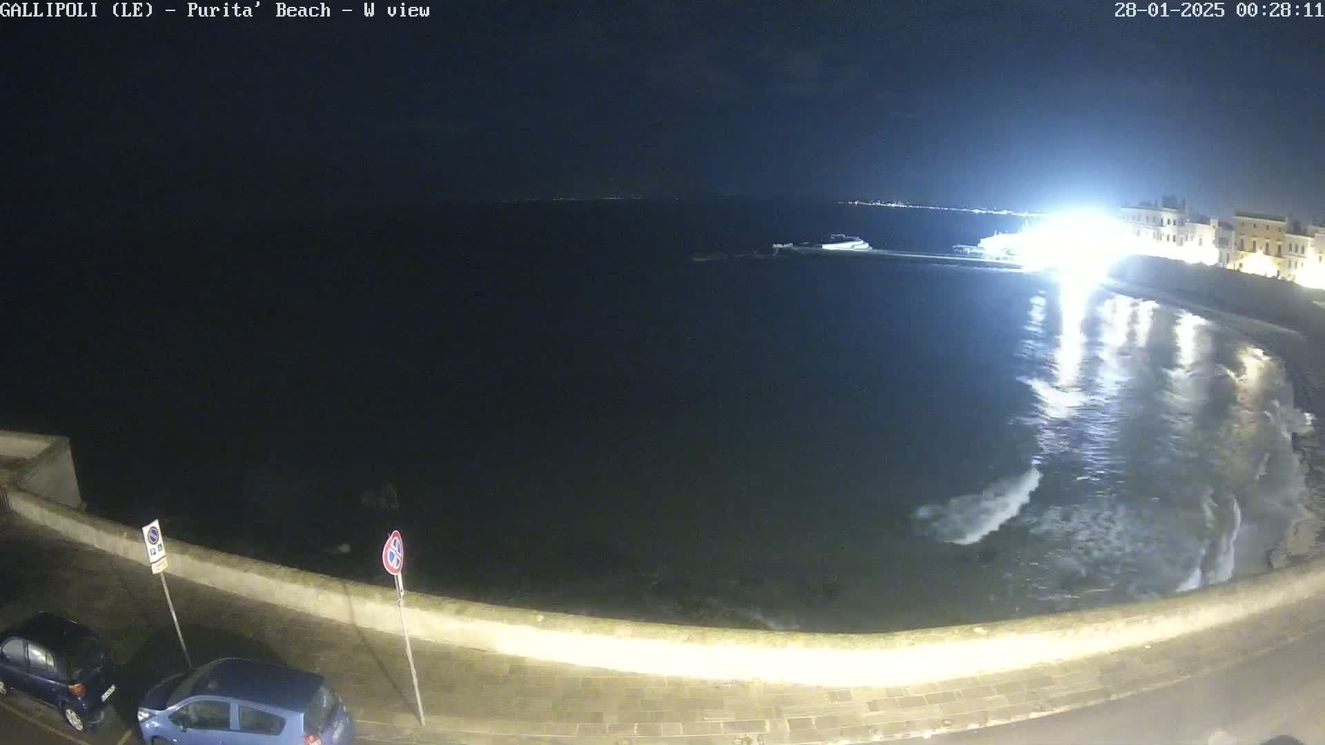 A nighttime view of a dark, wavy ocean meeting a low stone wall along a shoreline, with some cars parked nearby under a clear night sky.