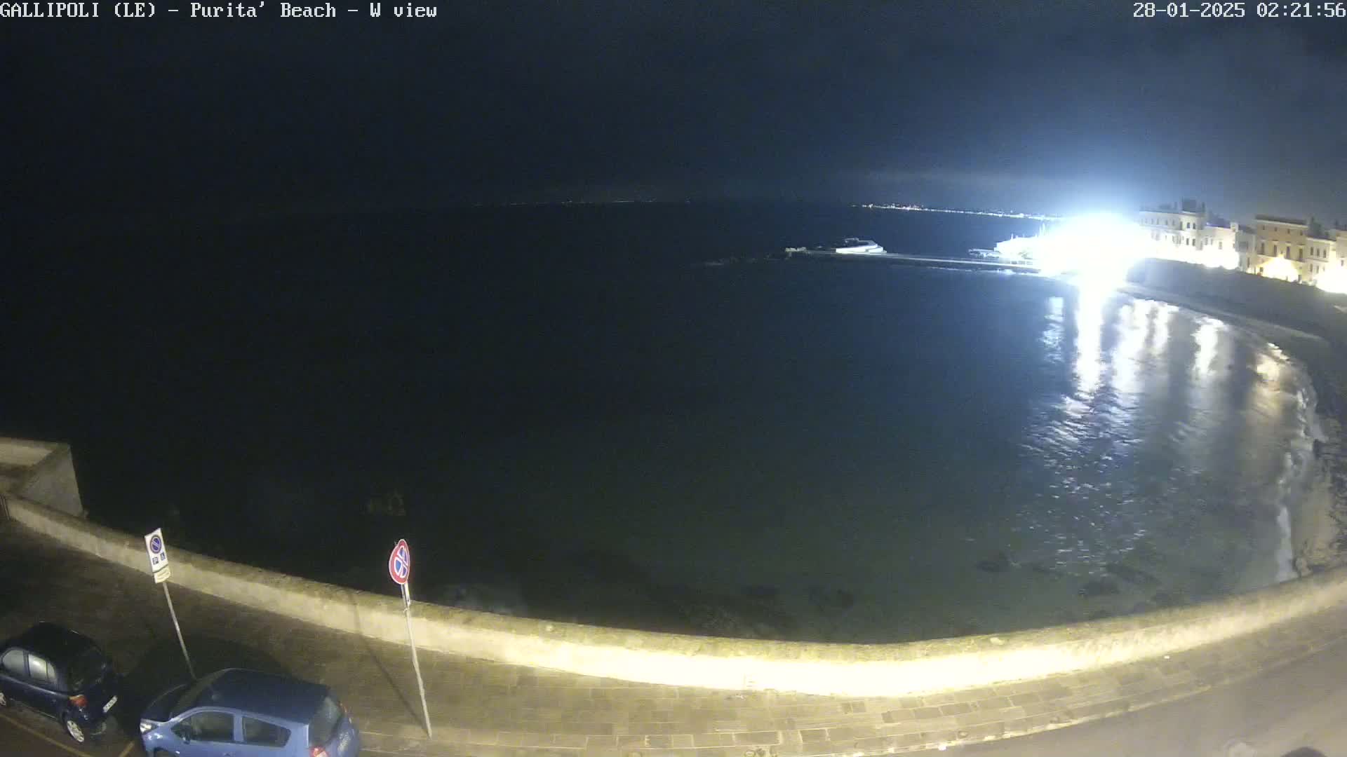 A nighttime view of a calm body of water next to a low stone wall with parked cars, under a dark sky.