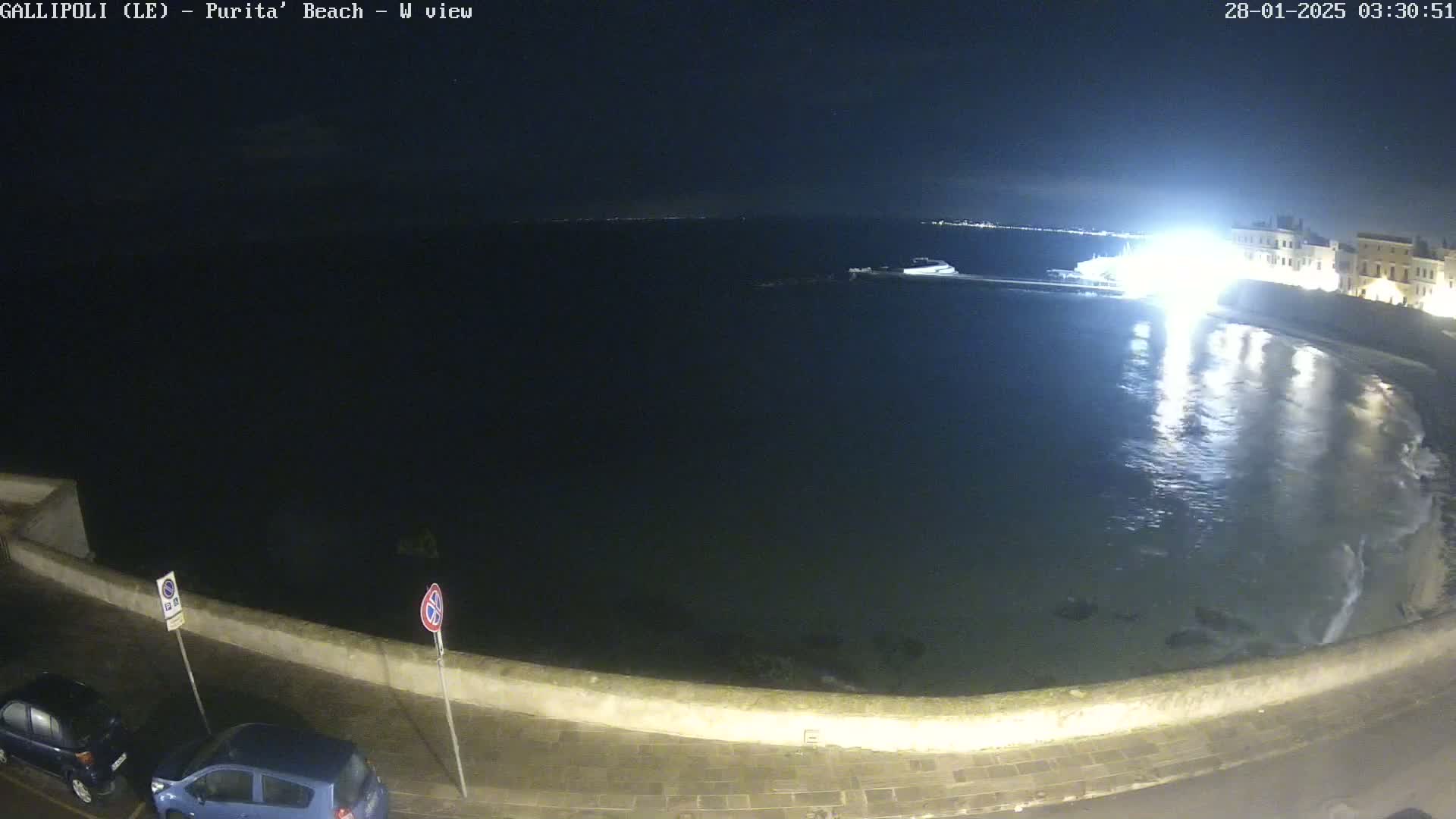 A nighttime view of a calm, dark body of water next to a low stone wall, with a few parked cars visible, under a clear, starry sky.