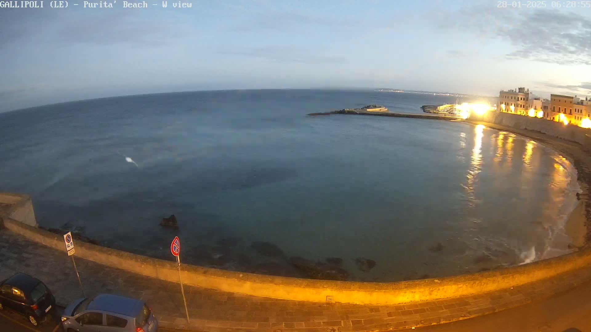 A calm, dark-blue sea laps gently against a low stone wall at twilight, with illuminated buildings visible in the background.