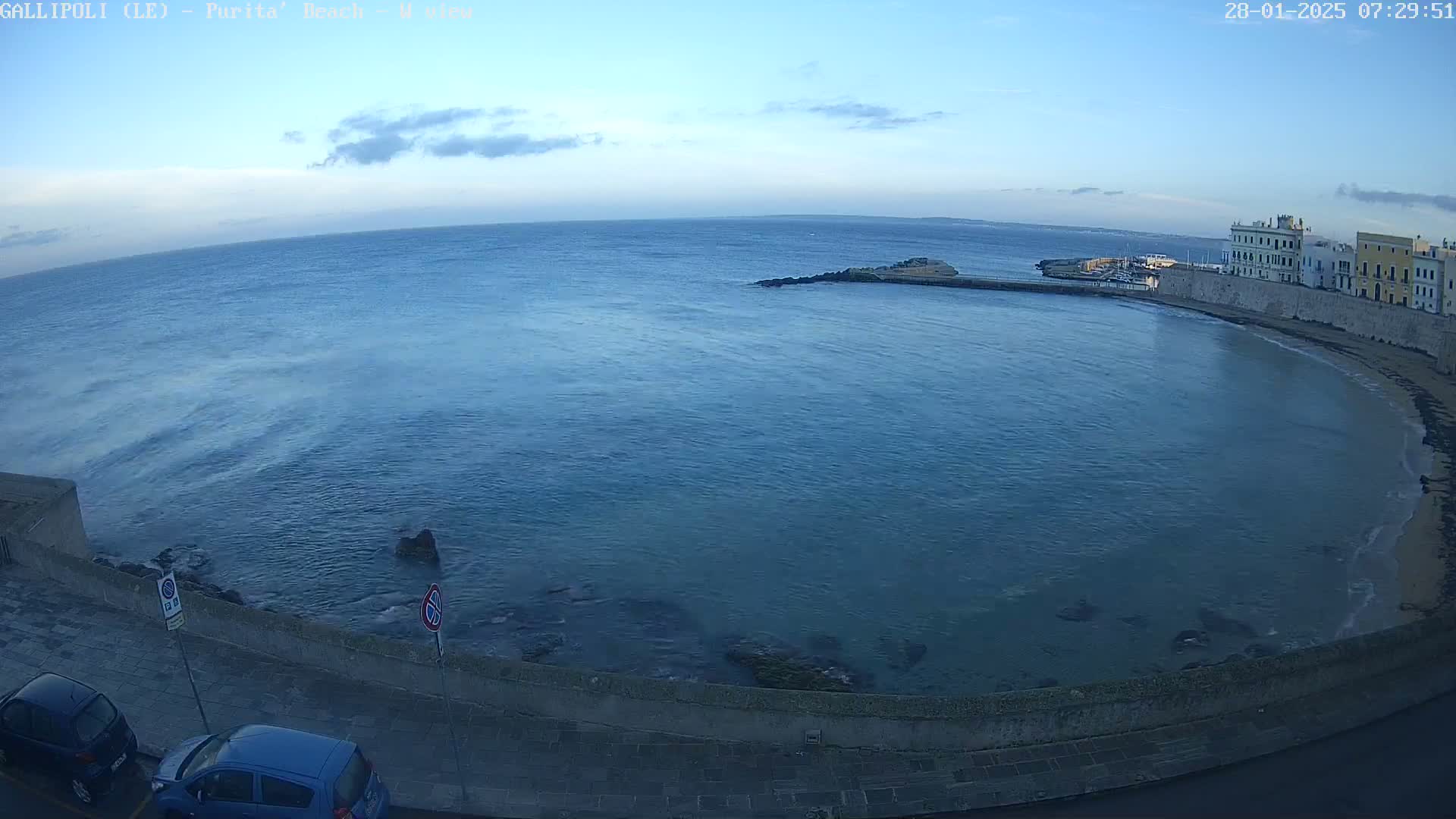 A partly cloudy, early morning view of a calm ocean bay curving around a seaside town with parked cars visible along a paved waterfront.
