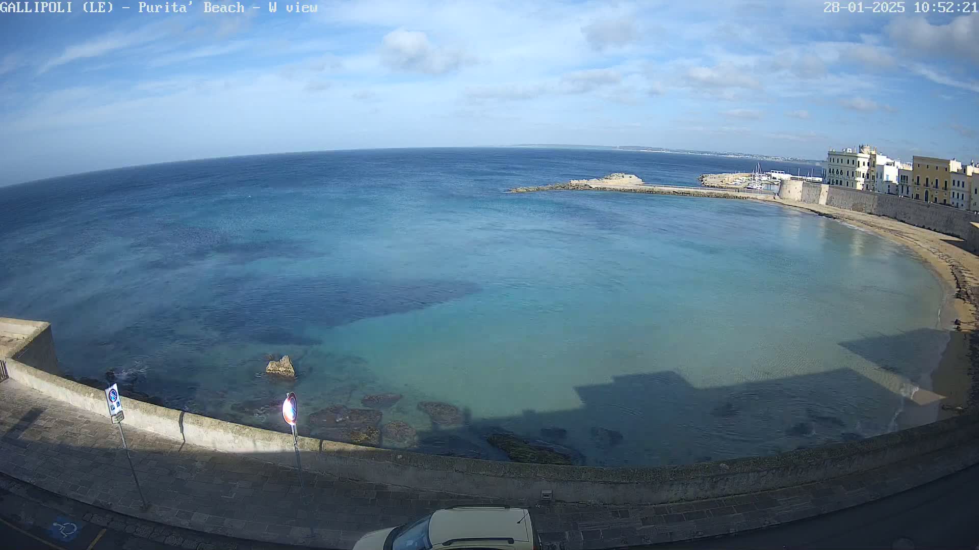 A partly sunny day shows a calm, clear, teal-colored bay with a sandy beach and stone buildings along a shoreline.