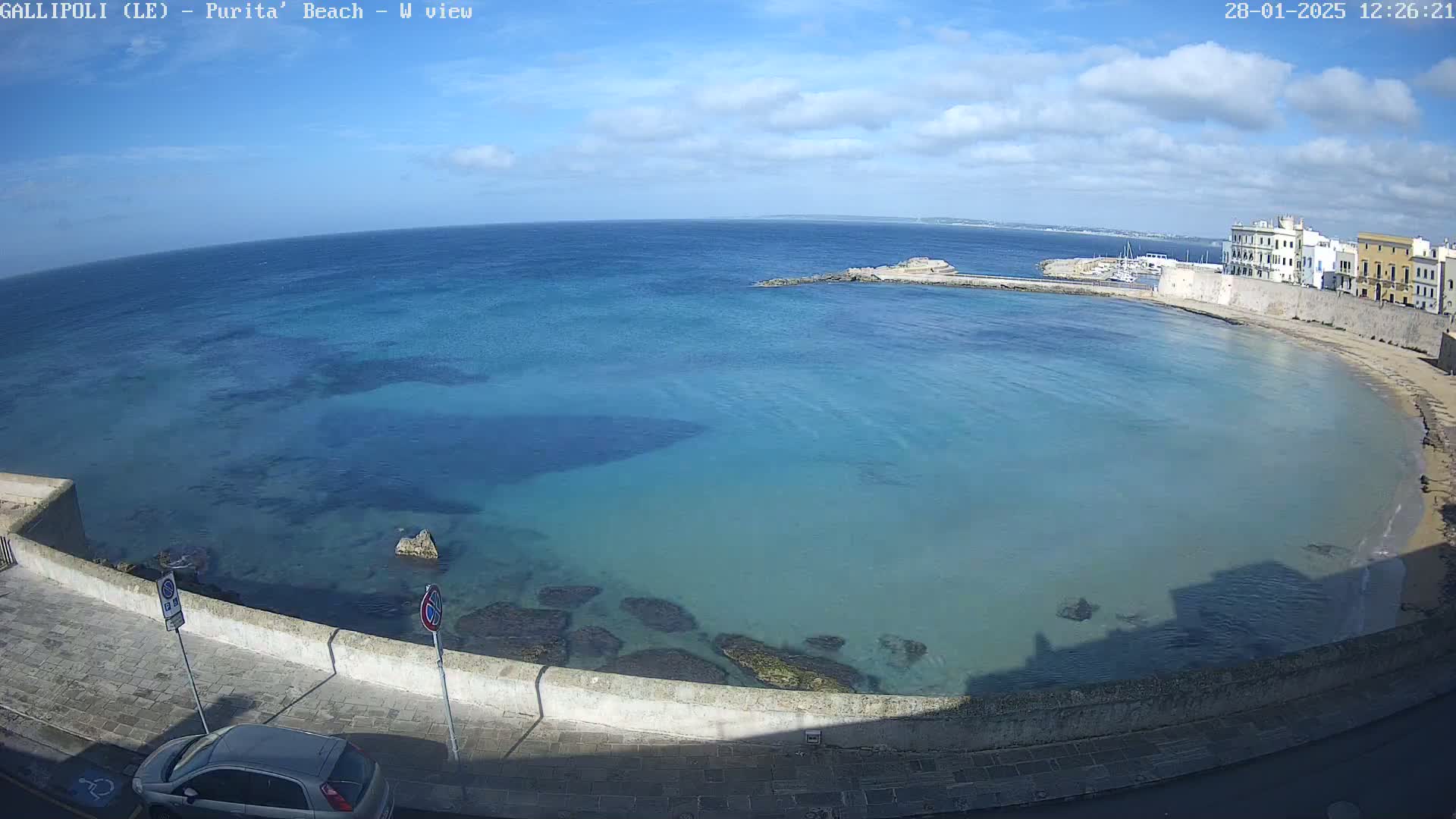 A partly cloudy sky overlooks a calm, clear turquoise bay with a stone wall and buildings along its edge.