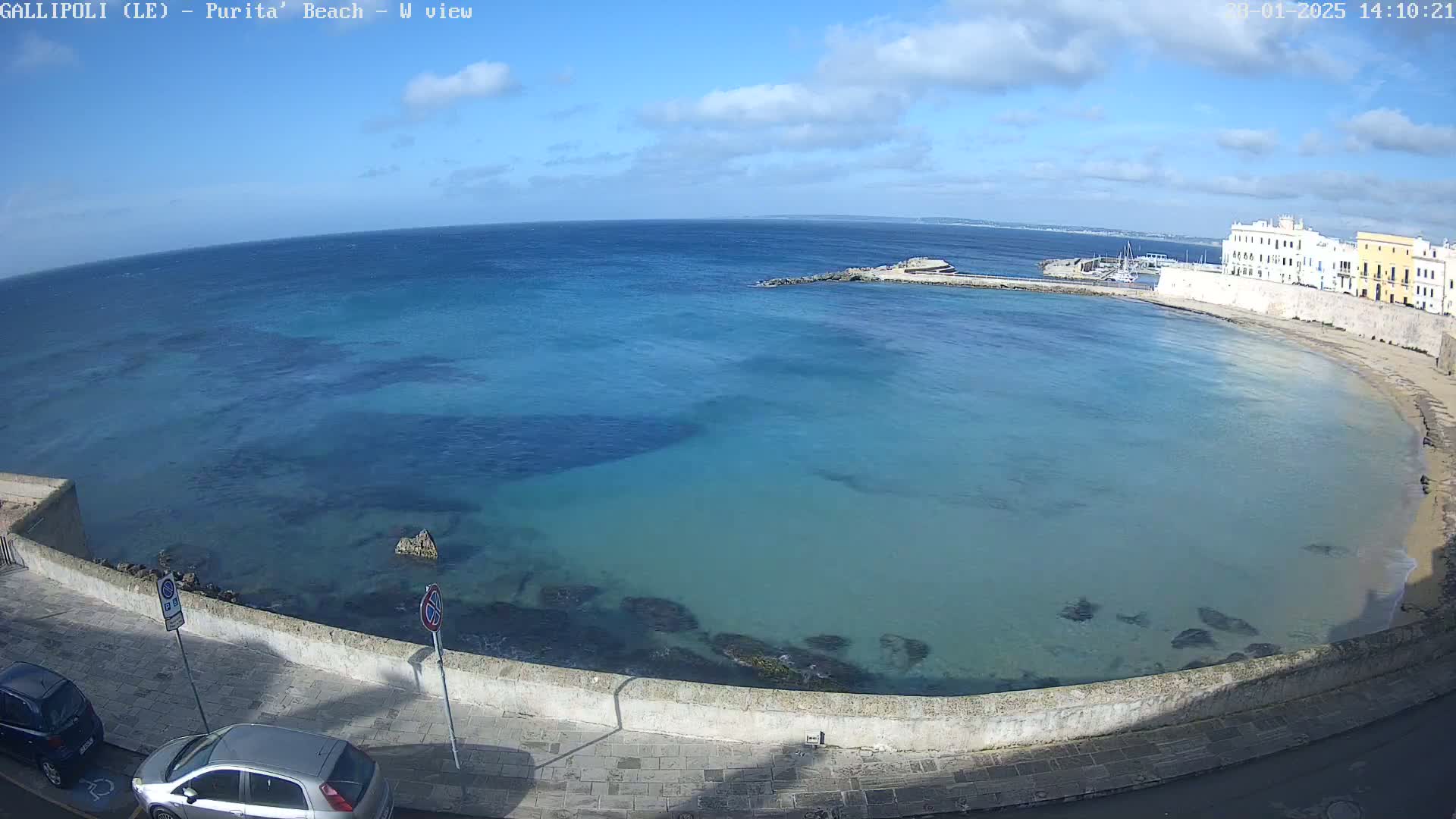 A clear, sunny day overlooks a calm, teal-colored bay curving around a stone wall, with buildings and a few parked cars visible.
