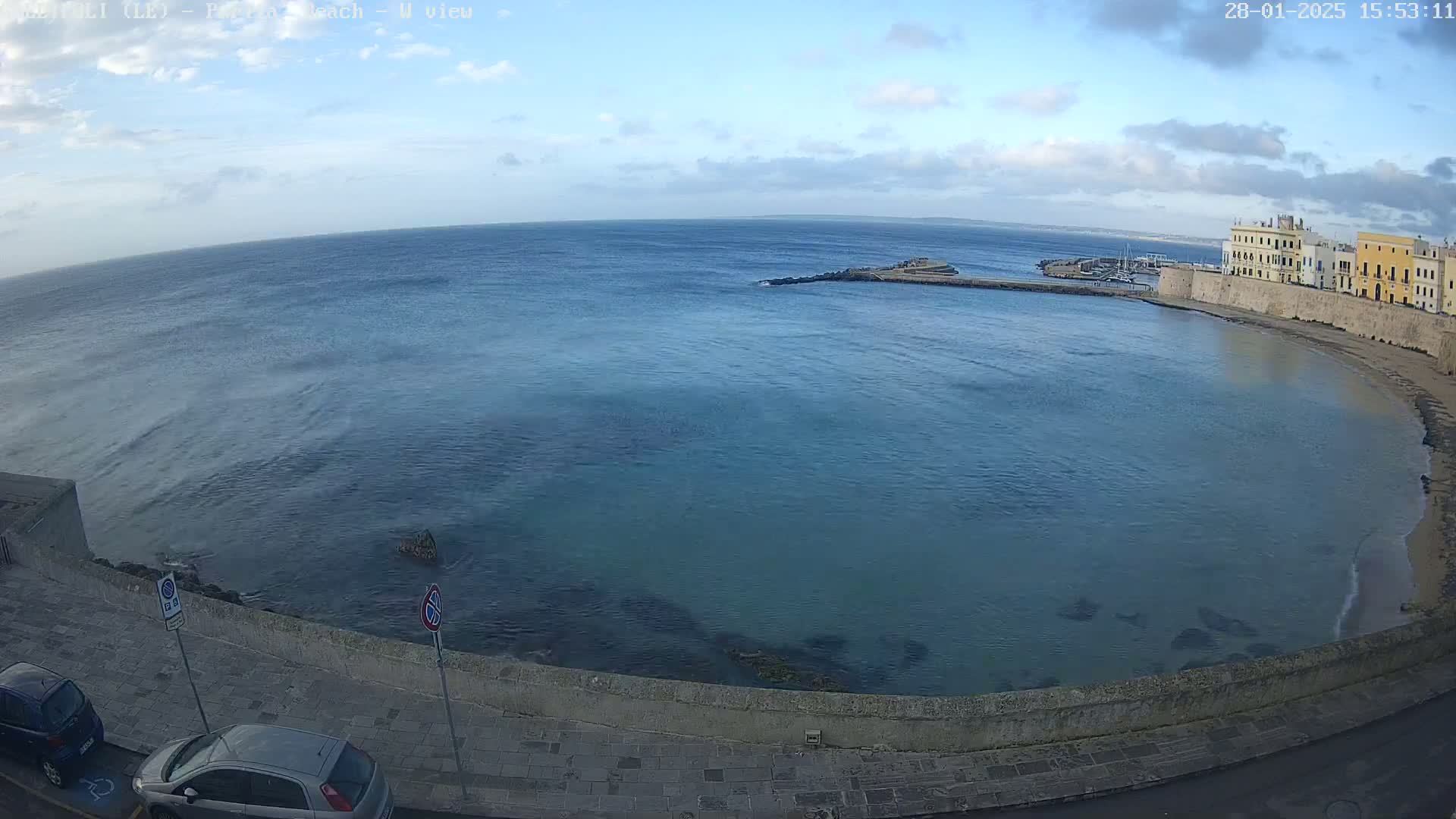 A partly cloudy day shows a calm sea curving around a stone wall and buildings.