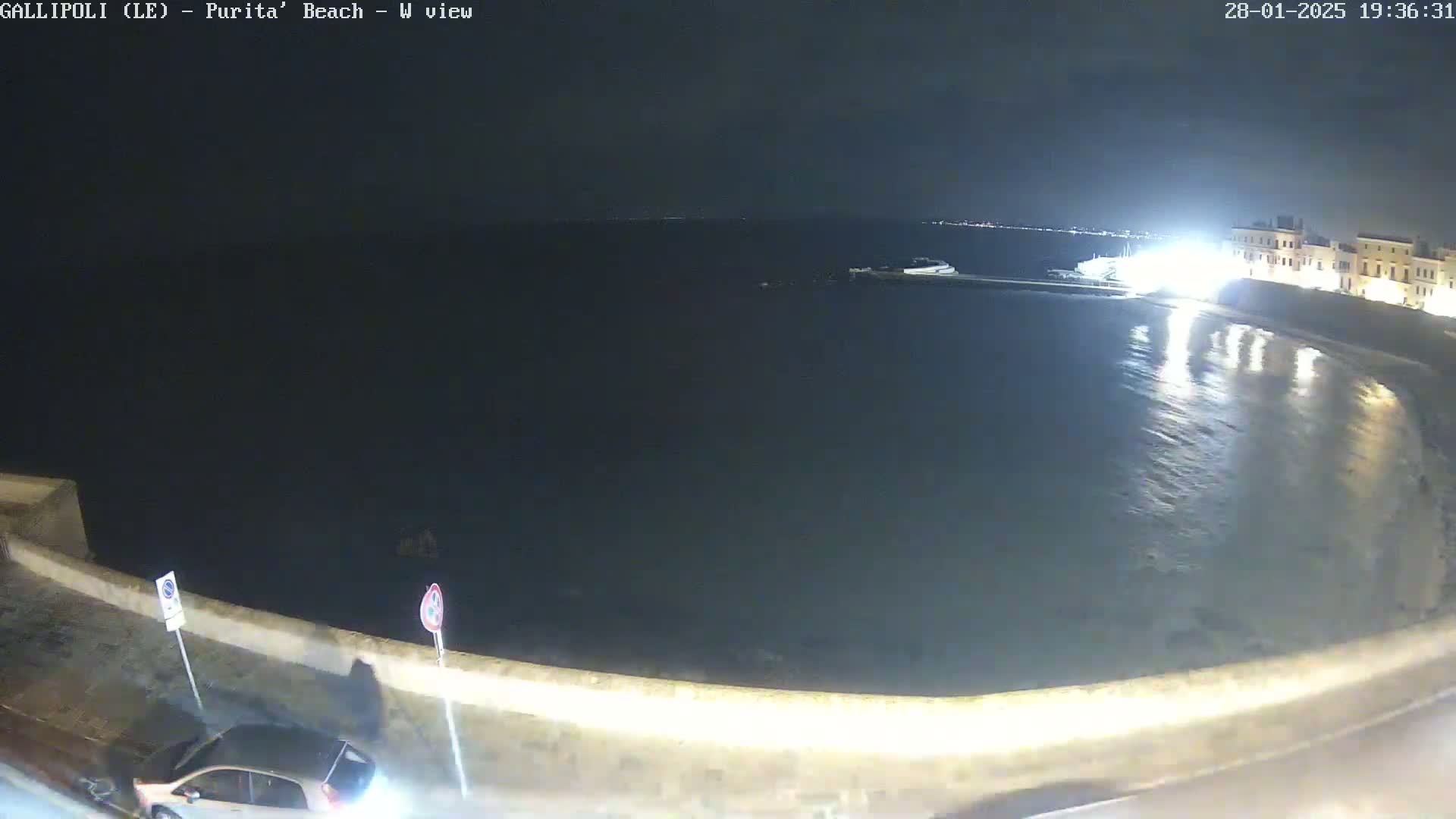 A nighttime, westward view of a calm, dark body of water next to a stone seawall and a town with illuminated buildings.