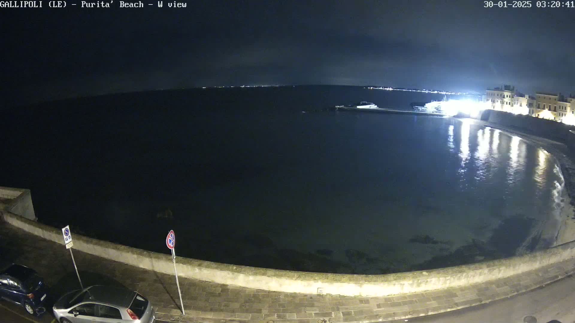 A nighttime view of a calm, dark body of water next to a stone wall and roadway, with a few cars parked nearby under a partly cloudy sky.