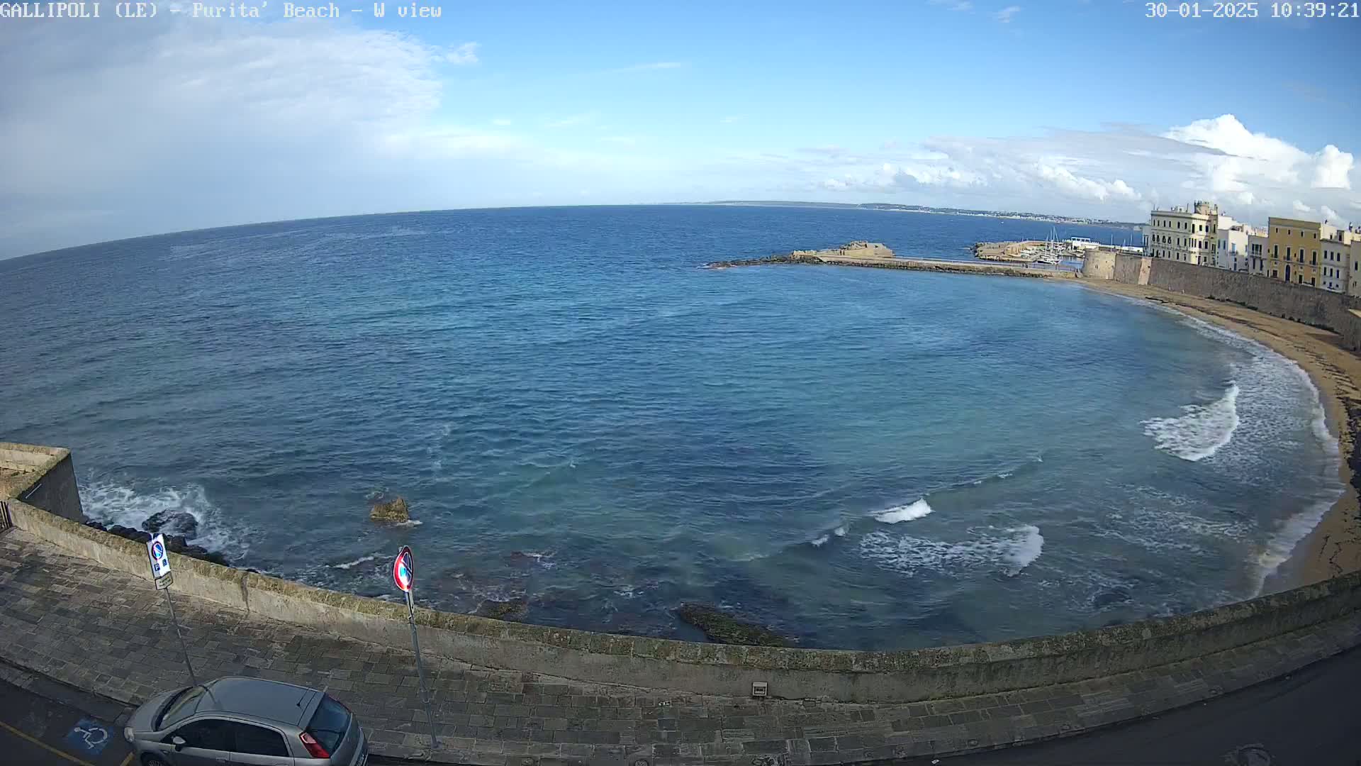 A mostly sunny day shows a curved bay with light waves lapping a sandy beach next to a stone wall and buildings, seen from an elevated position.