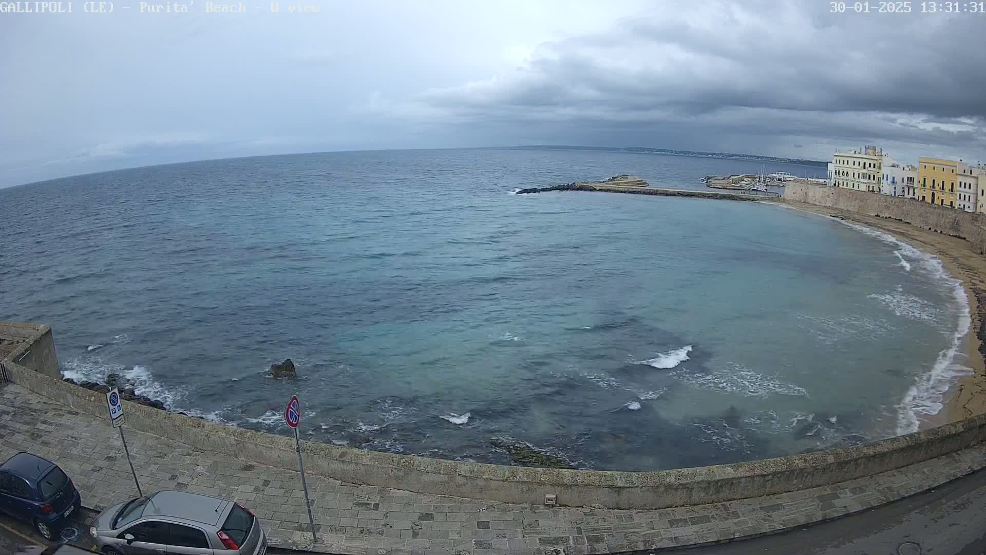 A partly cloudy day shows a coastal scene with a calm sea, a sandy beach, and buildings along a stone wall.
