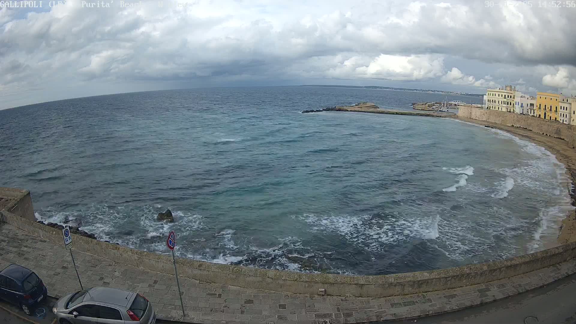 A partially cloudy day shows a rocky coast with small waves lapping a sandy beach and buildings beyond.