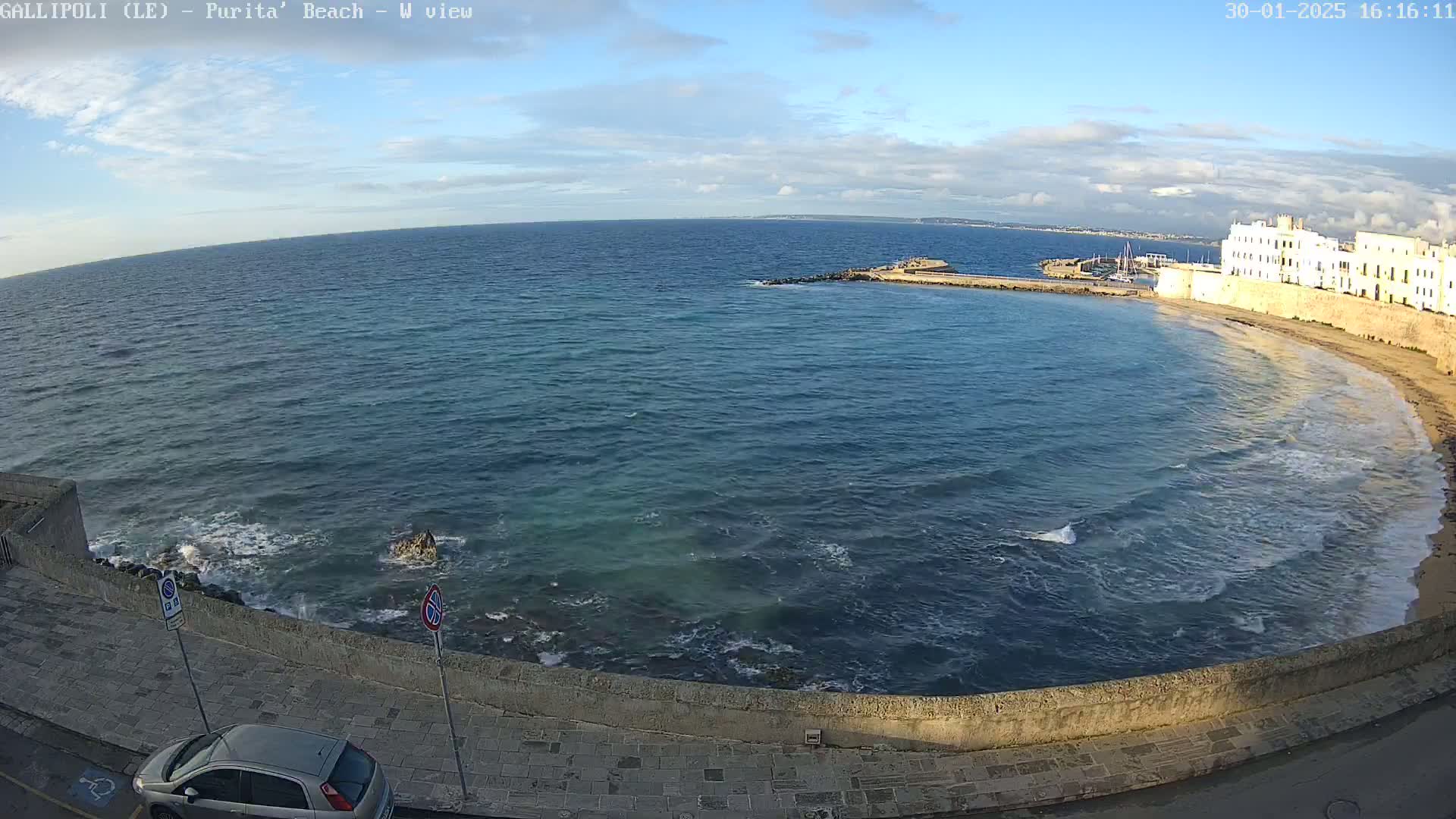 A partly cloudy day shows a view of a calm sea lapping a sandy beach next to a stone wall and buildings, with a car parked nearby.