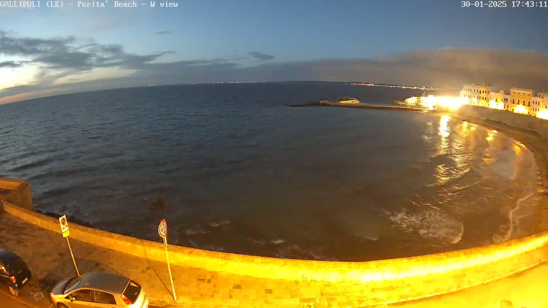 A nighttime view of a calm sea lapping gently against a low, illuminated seawall next to a roadway where two cars are parked.