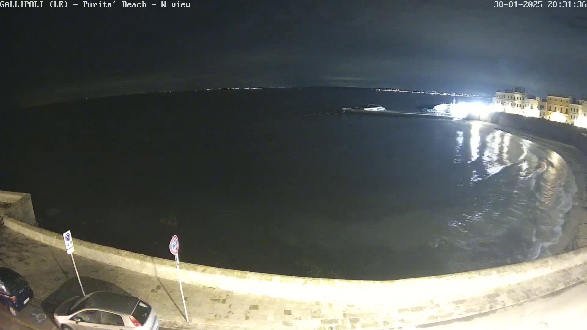A nighttime view of a calm, dark body of water lapping at a stone seawall, with a few lights visible across the water and some parked cars along the wall.