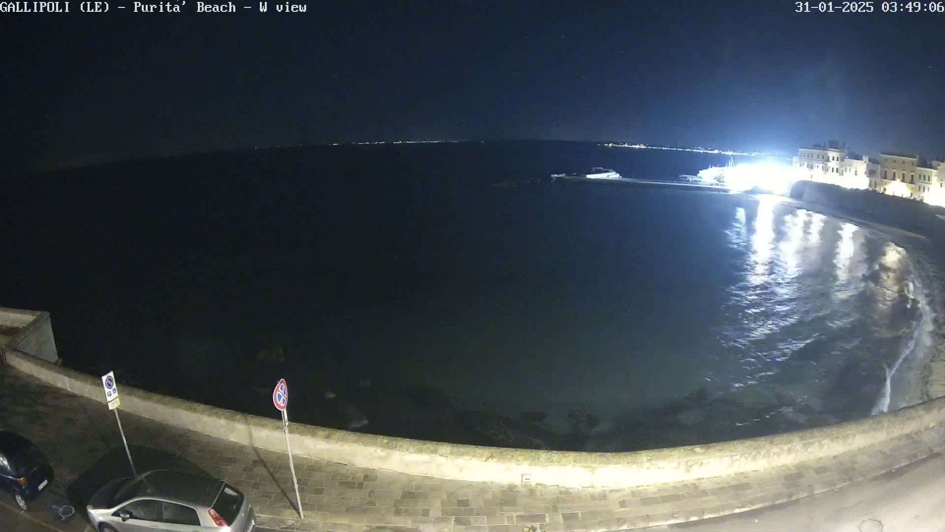 A nighttime view of a calm, dark sea reflecting lights from a nearby town, with a low stone wall and parked cars along a paved road in the foreground.