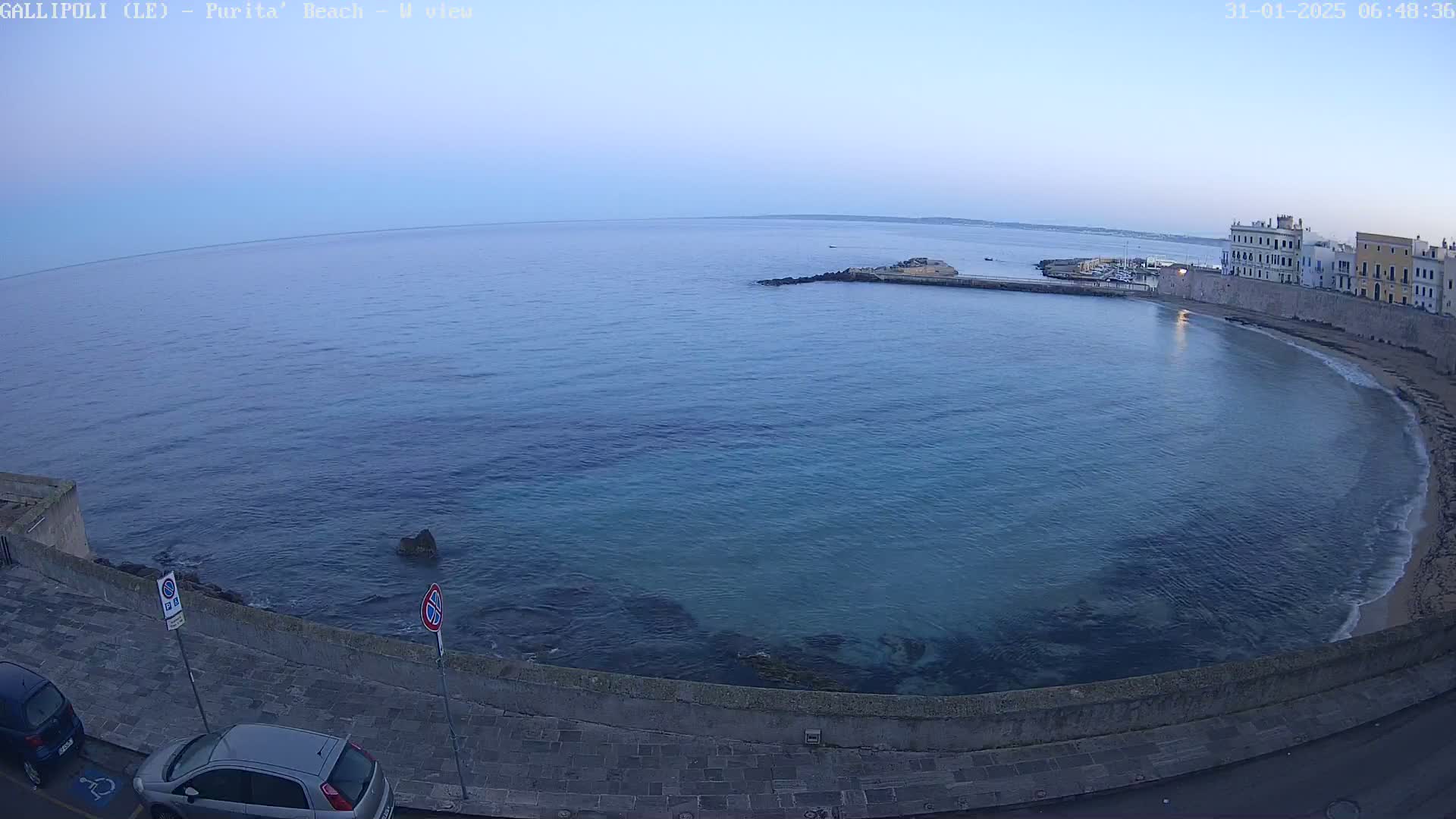 A calm, twilight-lit sea curves around a sandy beach and stone buildings, with a few cars parked along a paved road.