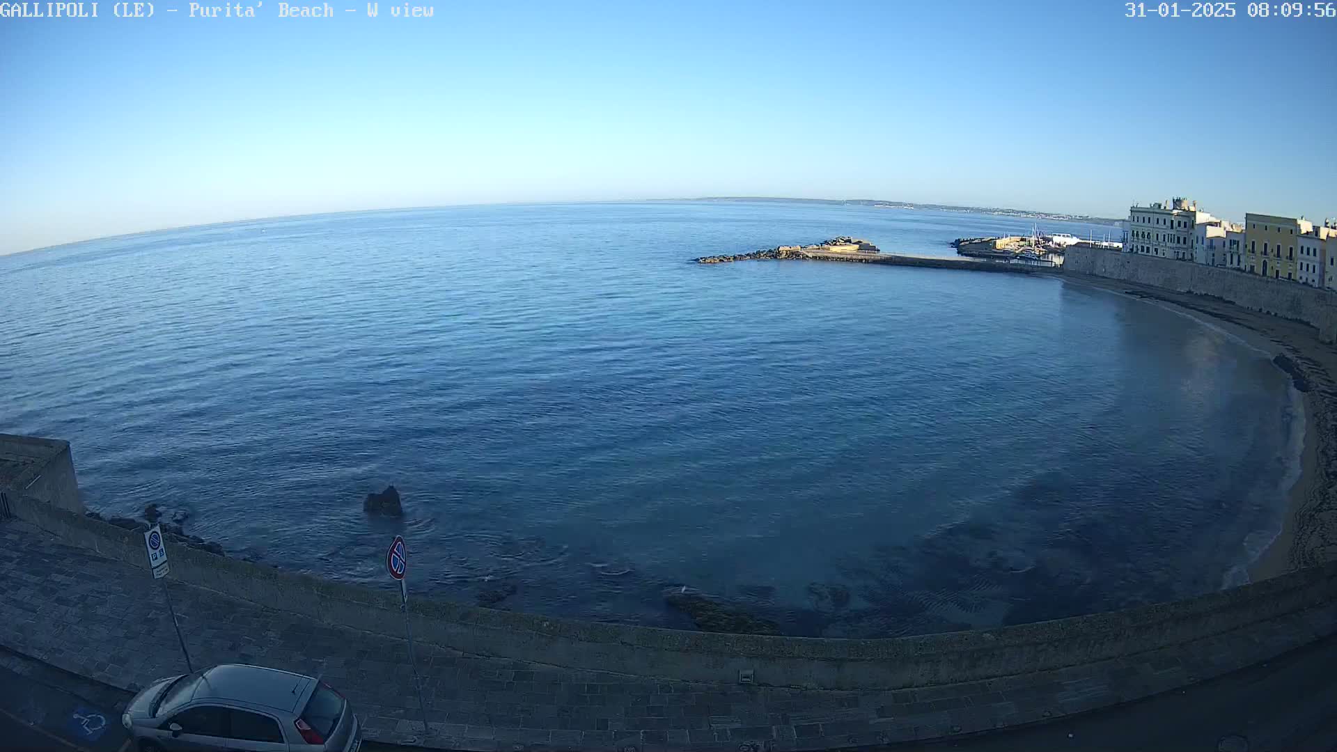 A clear, sunny day overlooks a calm blue ocean curving into a sandy beach next to a stone wall and buildings.