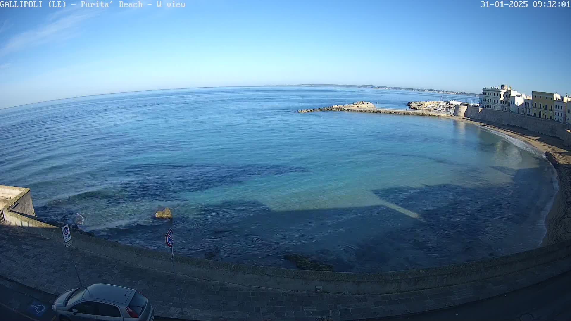 A clear, sunny day reveals a calm, blue sea lapping a sandy beach next to a stone wall and buildings.