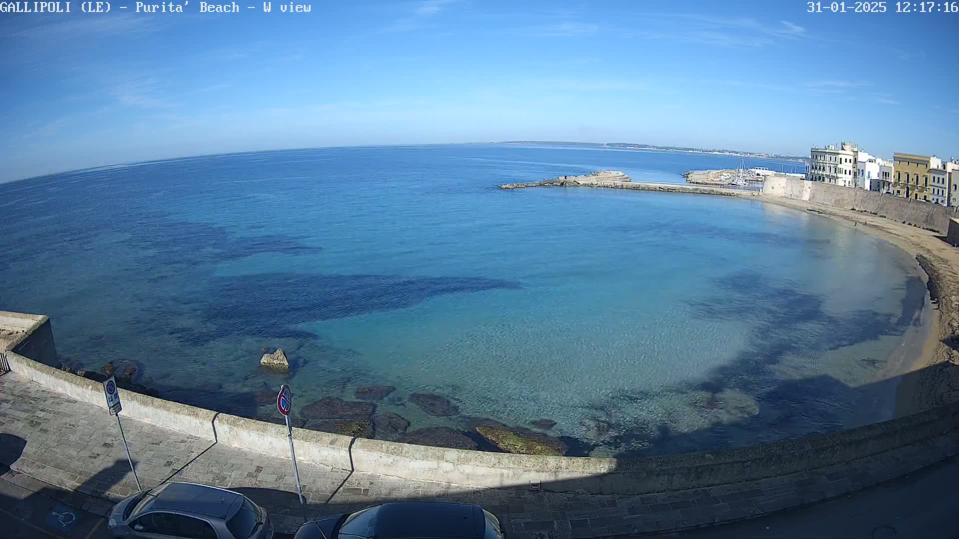 A clear, sunny day reveals a calm, turquoise bay curving around a sandy beach next to a stone-walled town.