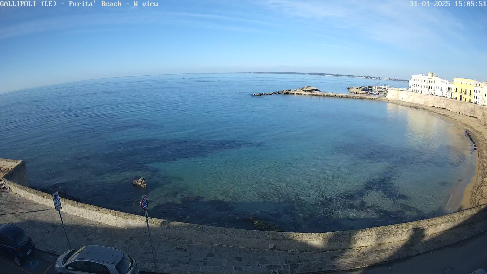 A clear, sunny day reveals a calm, blue-green sea curving into a sandy beach bordered by a stone wall and buildings.