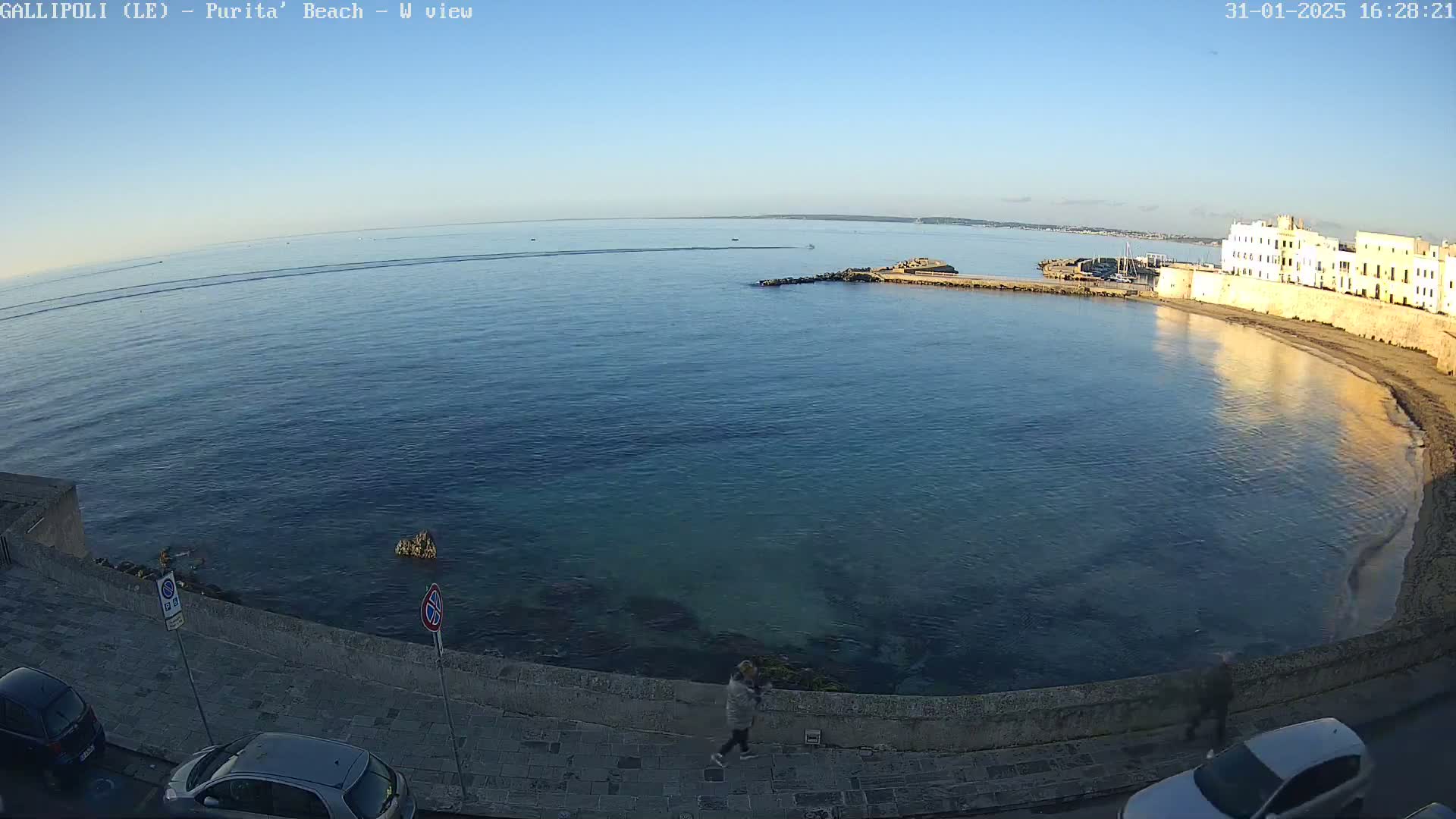 A clear, sunny day overlooks a calm bay with a stone wall and buildings lining the shore, a few people walking along the wall, and several parked cars visible.