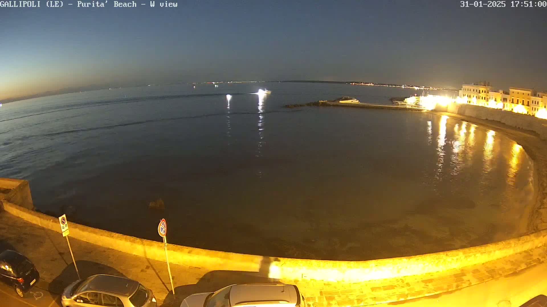 A nighttime view of a calm, dark-blue sea curving into a crescent-shaped bay, illuminated by lights along a stone wall and buildings bordering the shore.