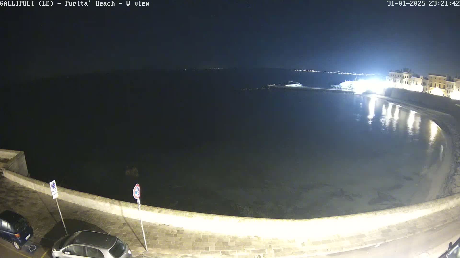 A nighttime view of a calm, dark body of water next to a low-lit stone wall with cars parked nearby.