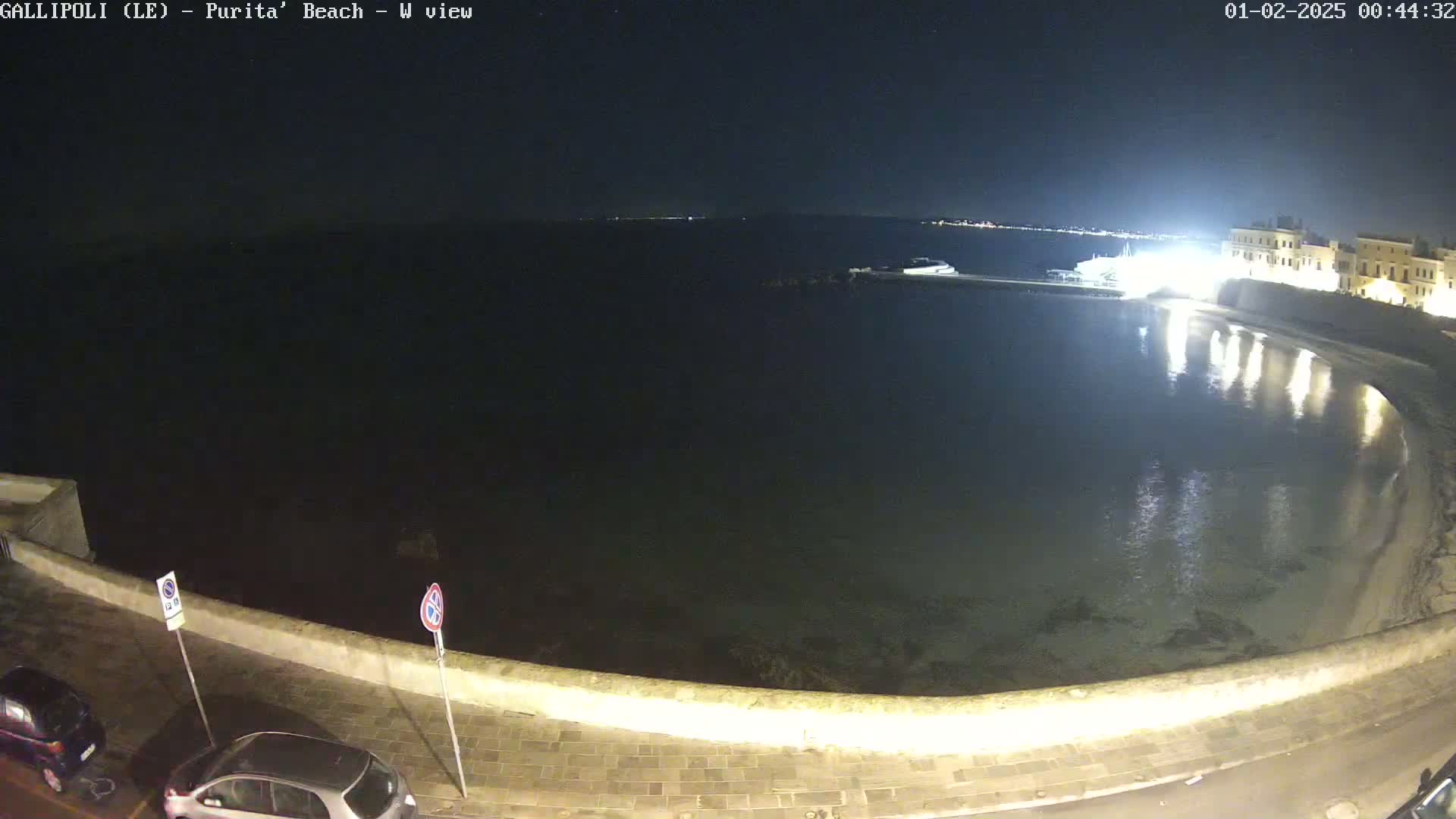 A nighttime view of a calm, dark body of water curving along a low stone wall, with a few parked cars visible on the land next to the water.