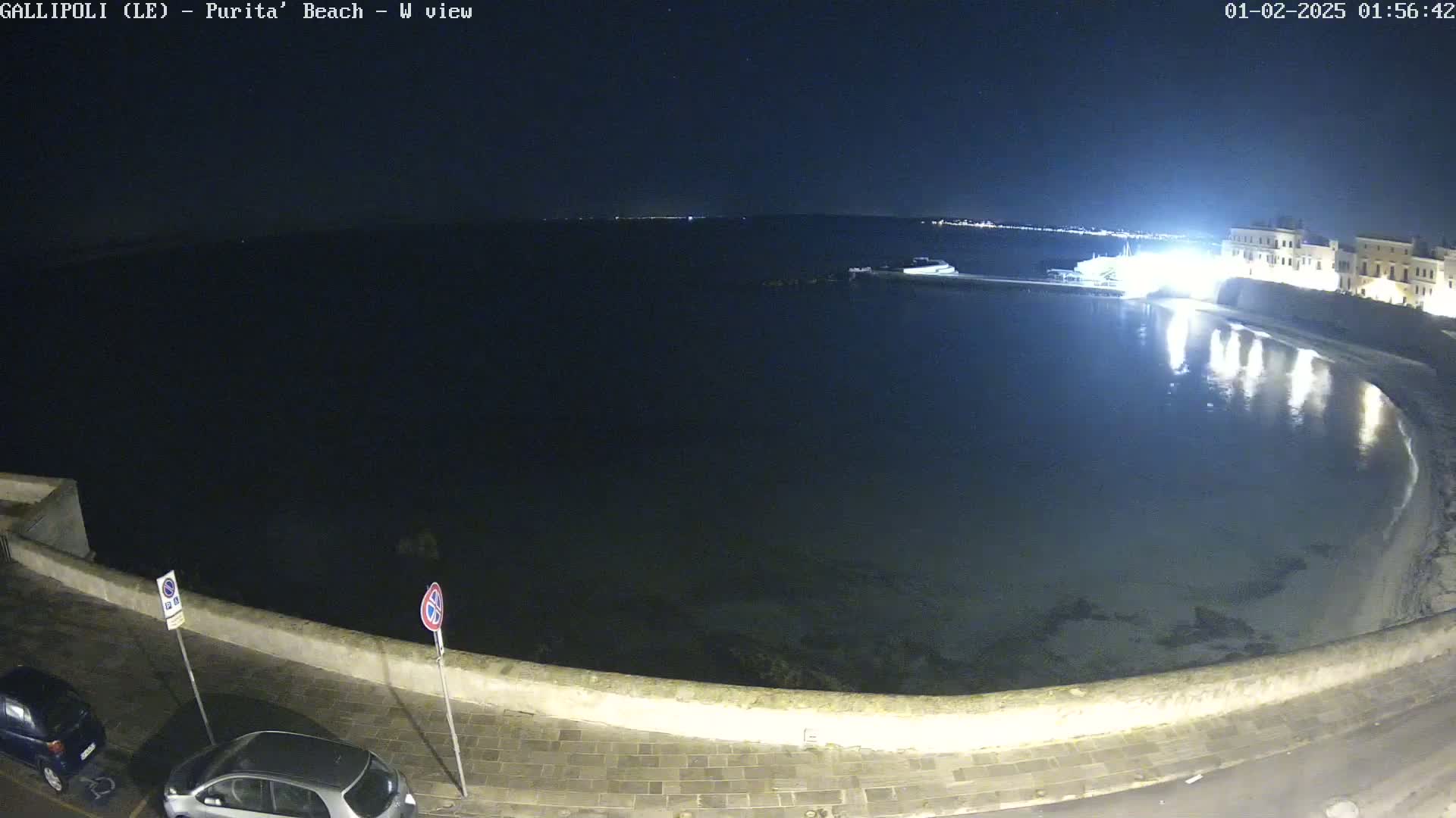 A nighttime view of a calm, dark-blue body of water next to a stone wall, with a few parked cars visible and a building illuminated in the background.