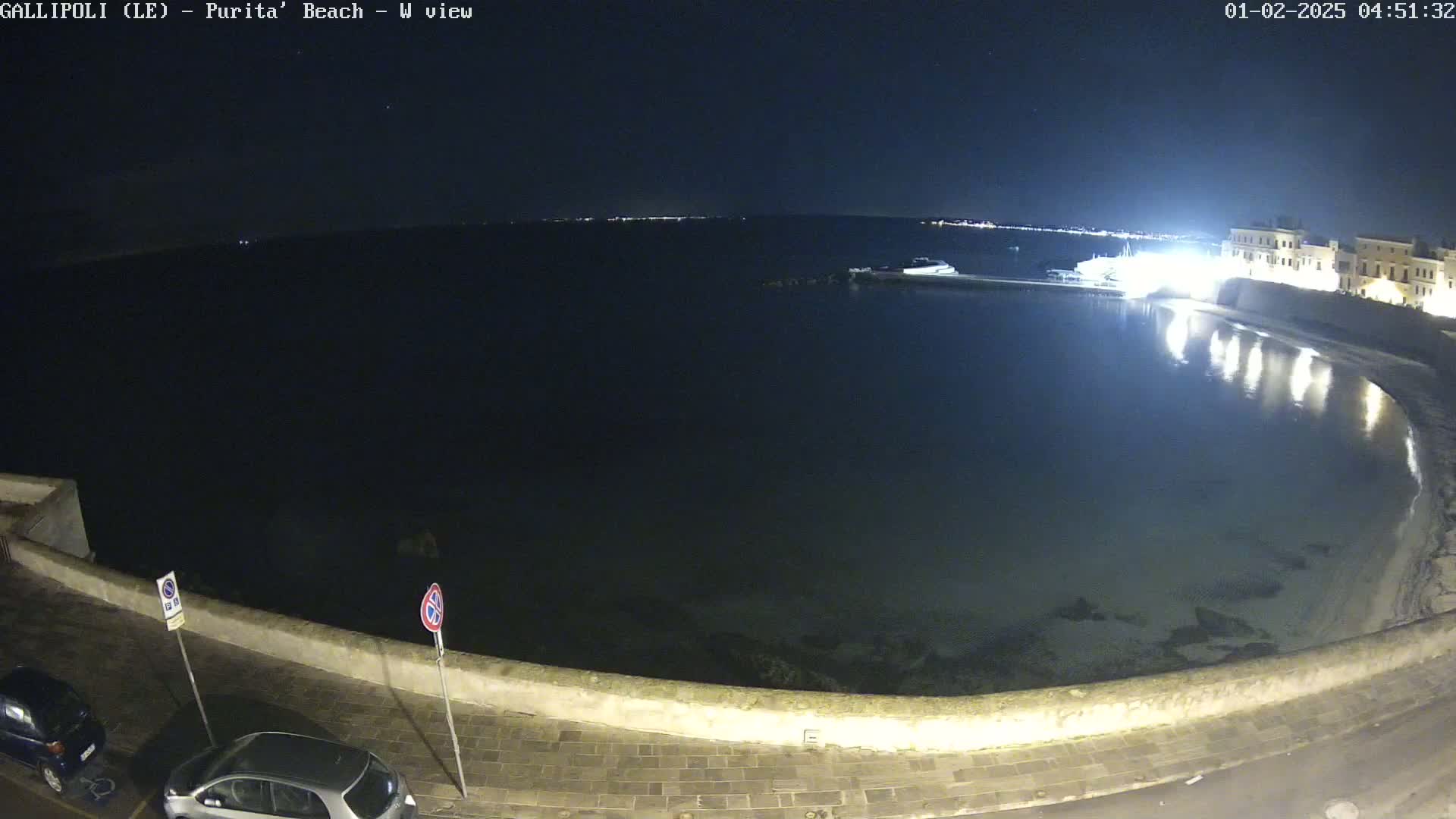 A nighttime view of a calm bay, illuminated buildings, and parked cars along a stone wall.