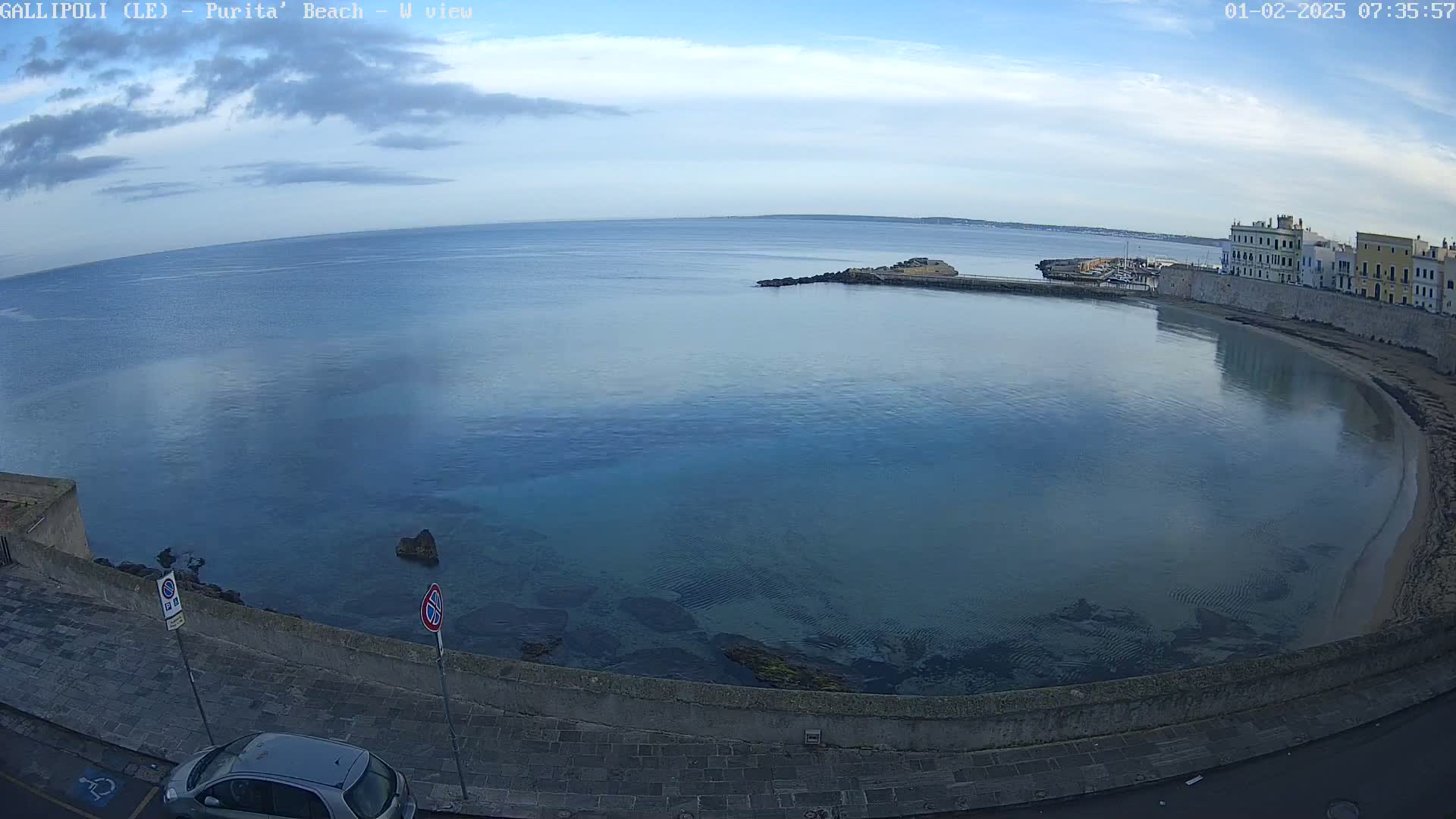 A calm, clear body of water curves around a stone embankment, with buildings visible in the distance under a partly cloudy sky.