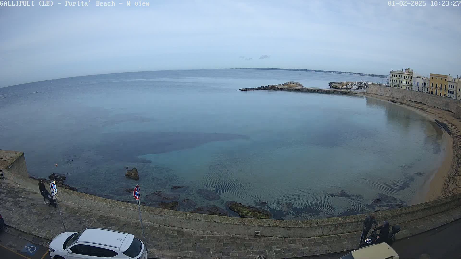 A calm, overcast day reveals a curved bay with clear, shallow turquoise water lapping a stone shoreline, adjacent to a paved walkway and buildings.