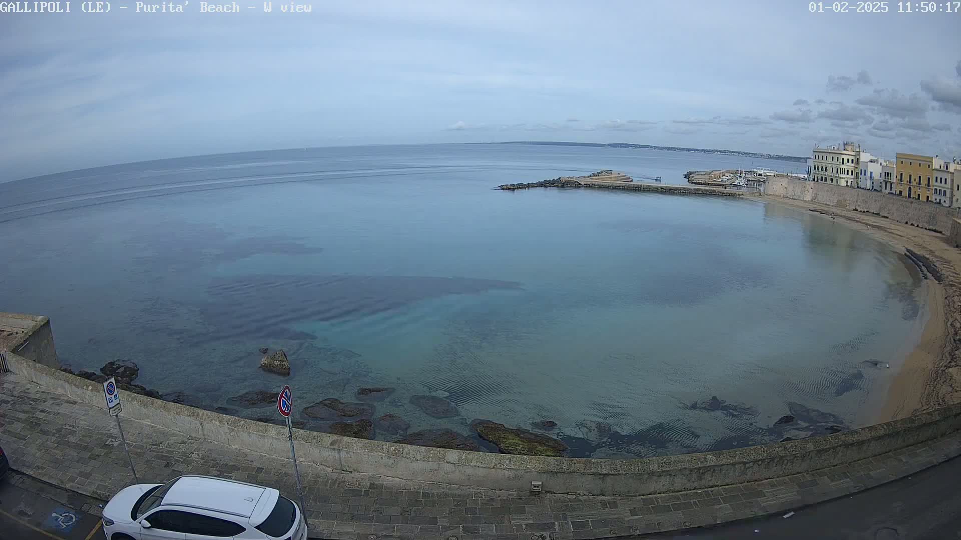 A tranquil, clear bay with a sandy beach curves around a stone wall and buildings under a mostly cloudy sky.