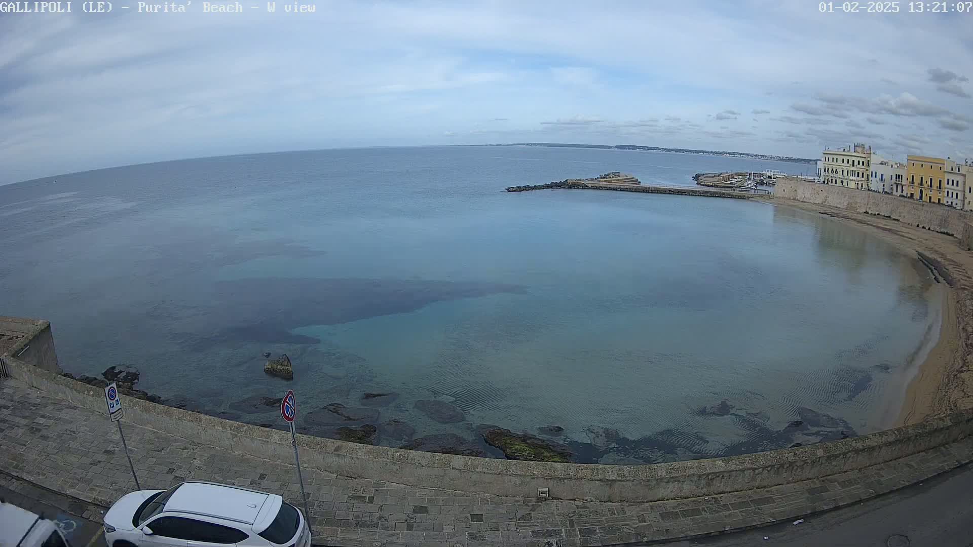 A mostly clear day shows a calm, shallow bay with a sandy beach and buildings along the shoreline.