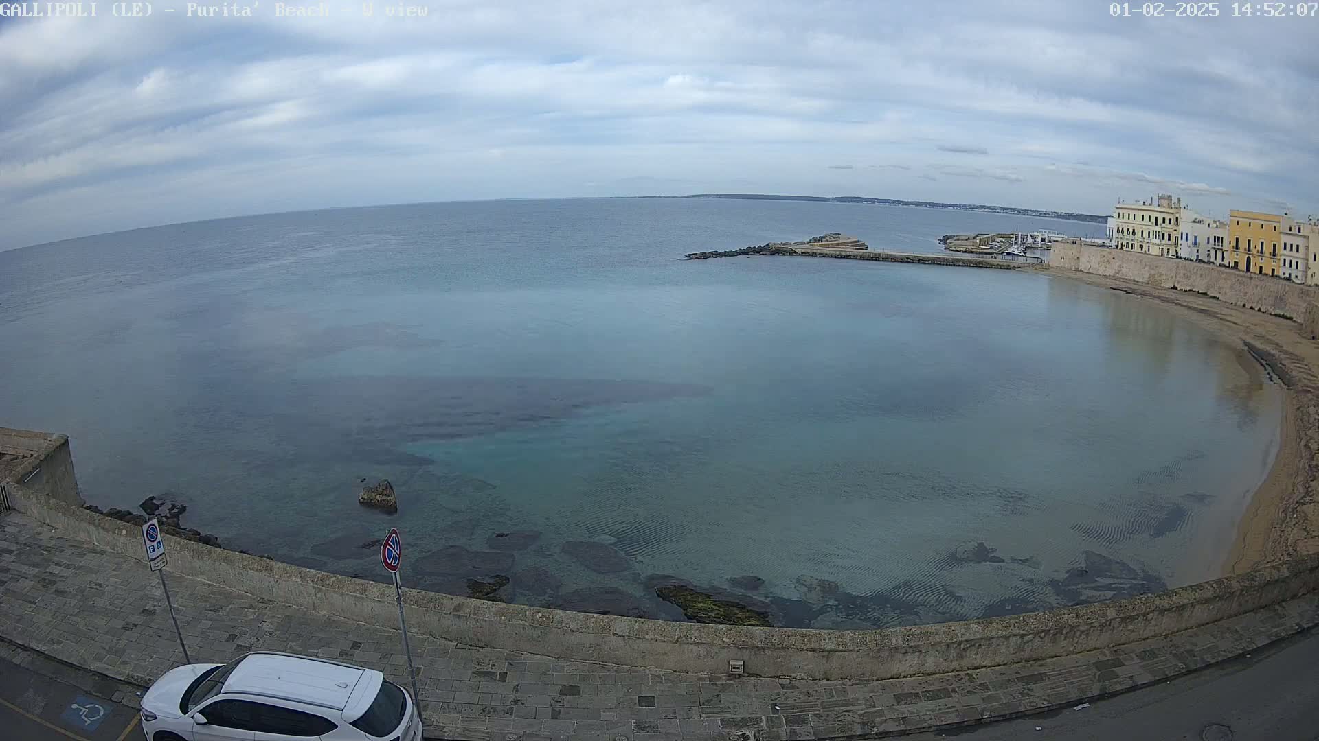 A partly cloudy day reveals a tranquil bay with clear, shallow water, a sandy beach, and buildings lining the shore.