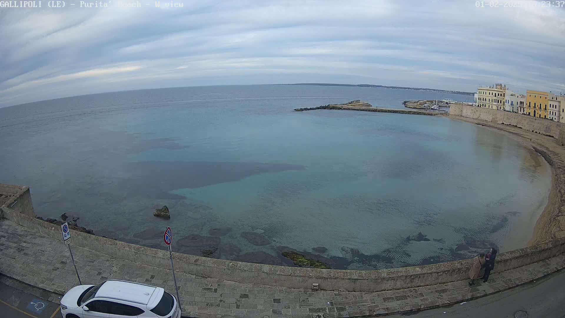 A coastal scene under a cloudy sky shows a calm, clear, teal-colored bay curving around a stone embankment with buildings and a white car parked along the street.