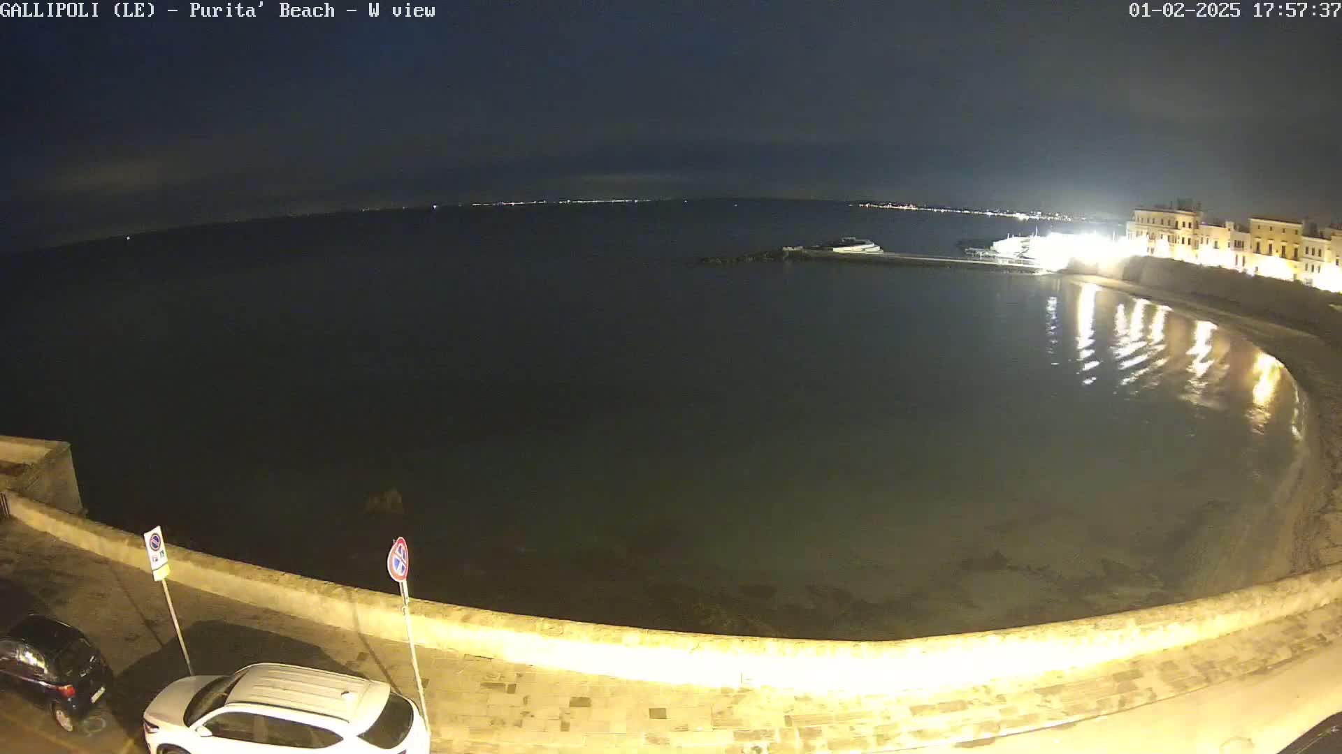 A nighttime view of a calm body of water next to a stone wall and buildings, with a few cars parked nearby under a dark sky.
