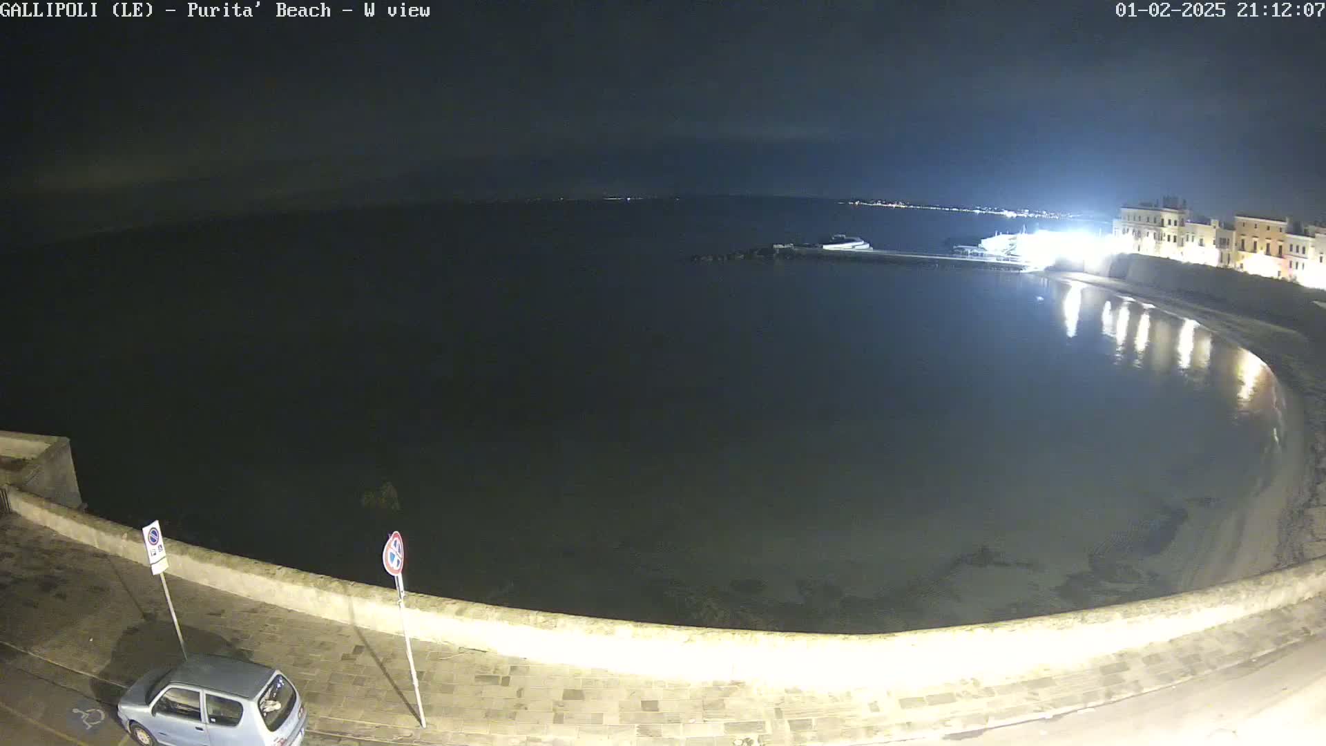 A nighttime, high-angle view shows a calm, dark body of water next to a paved waterfront area with a single car parked nearby.