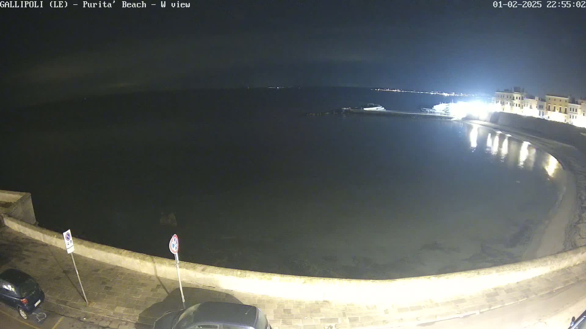 A nighttime view of a calm, dark body of water next to a low stone wall, with buildings and parked cars visible along the shore.