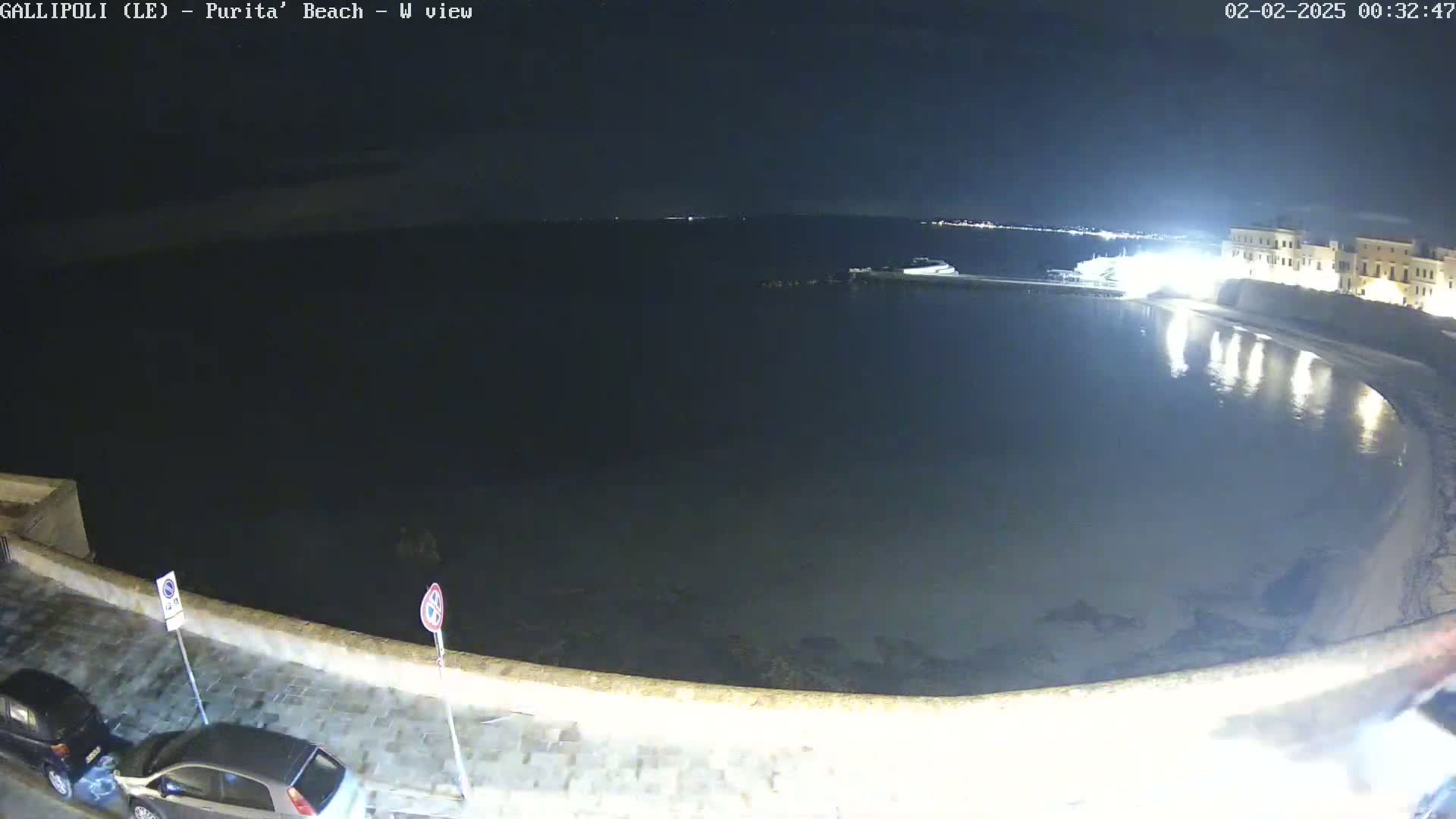 A nighttime view of a calm, dark body of water next to a stone wall and buildings, with a few cars parked along the wall.