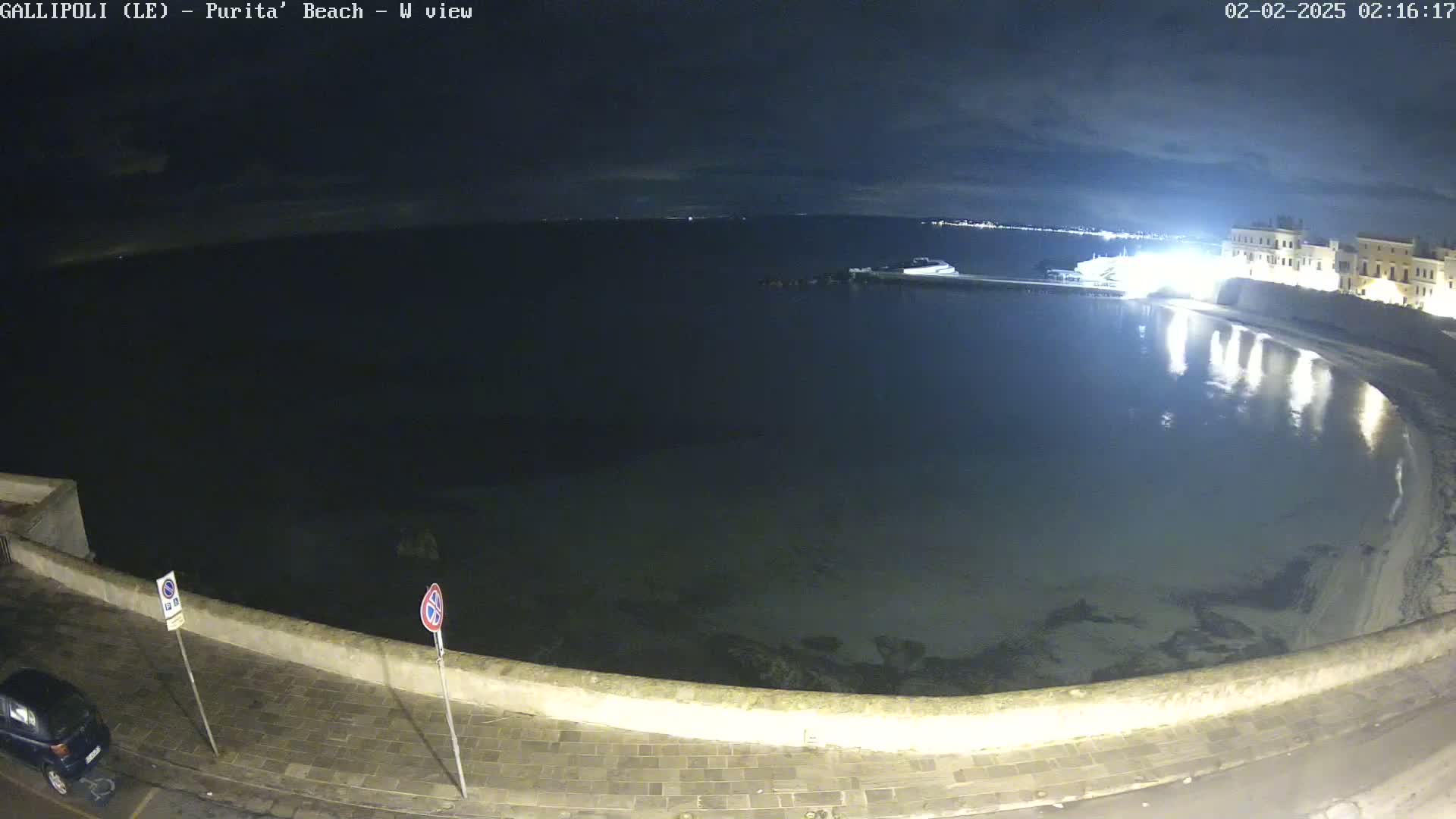 A nighttime, seaside view shows a calm body of water reflecting lights from buildings on the shore under a partly cloudy sky.