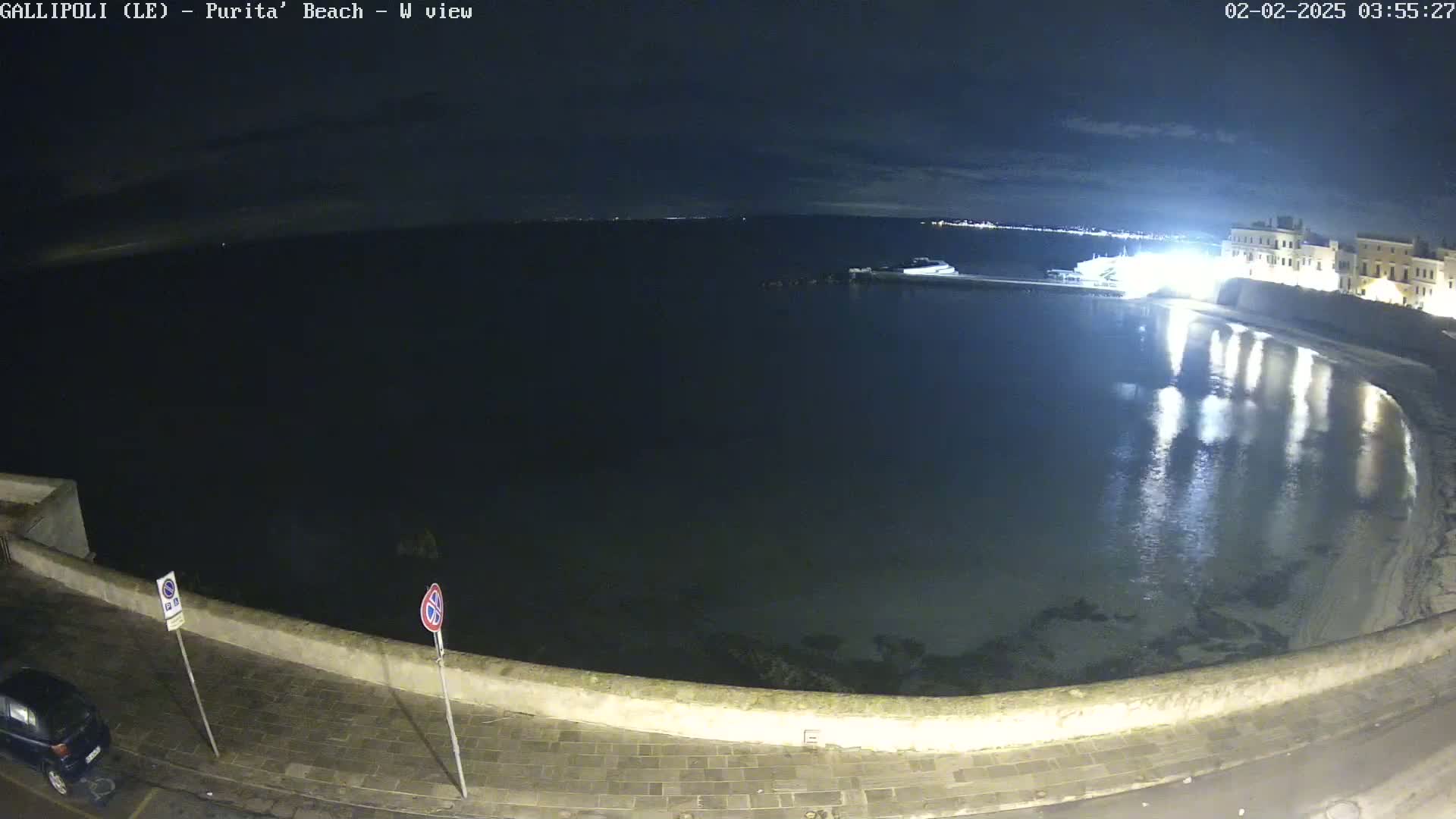 A nighttime view of a calm, dark body of water reflecting nearby lights, next to a stone wall and paved area with a car parked.
