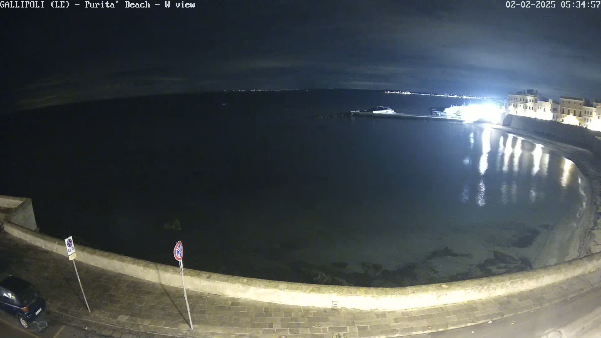 A nighttime view of a calm, dark body of water next to a low stone wall and buildings, under a mostly clear sky.