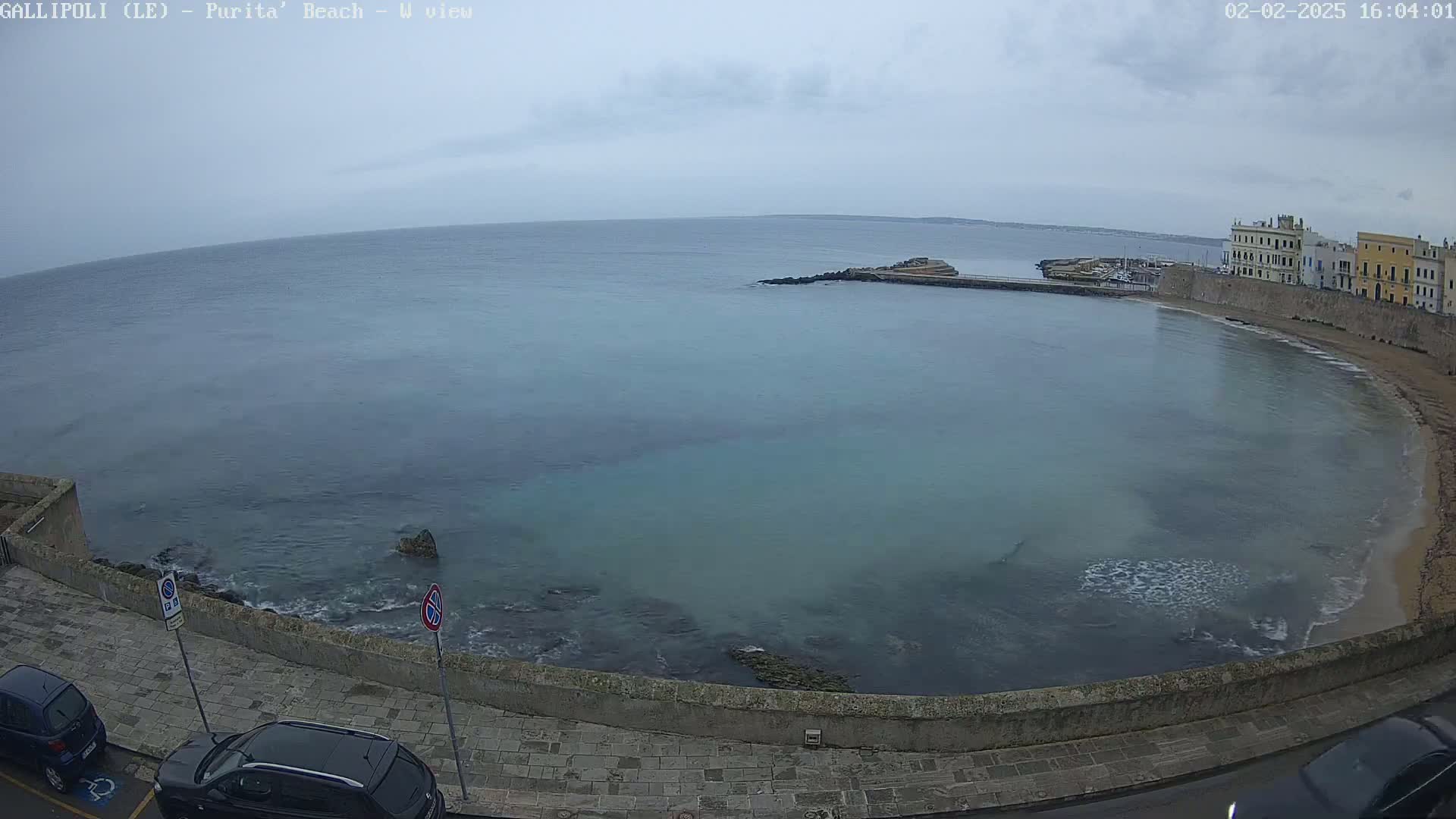 A calm, overcast day shows a curved bay with a stone wall and buildings along the shore, several cars parked nearby, and a partially visible sandy beach.