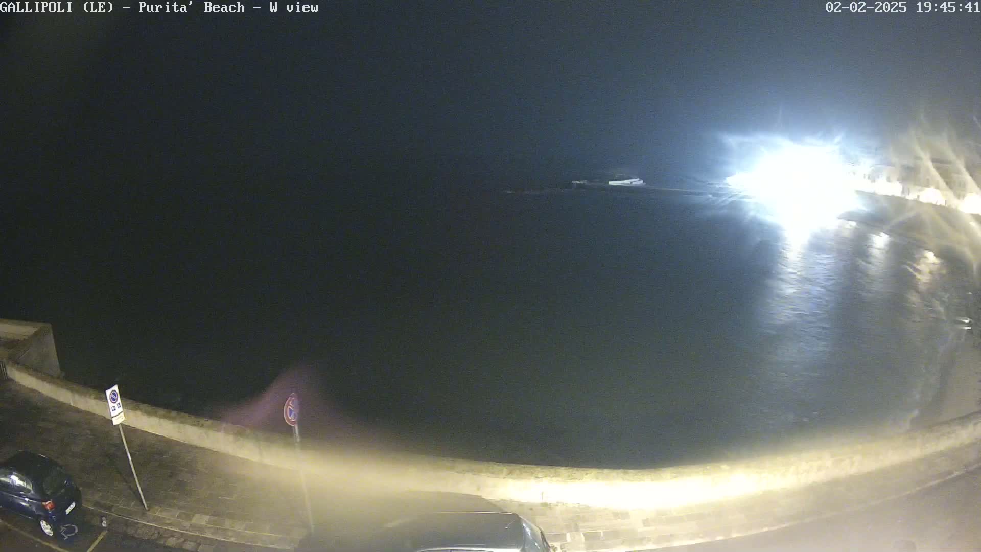 A nighttime, seaside view shows a dark body of water reflecting bright lights from a nearby shoreline, with a low wall and parked cars visible along the road.