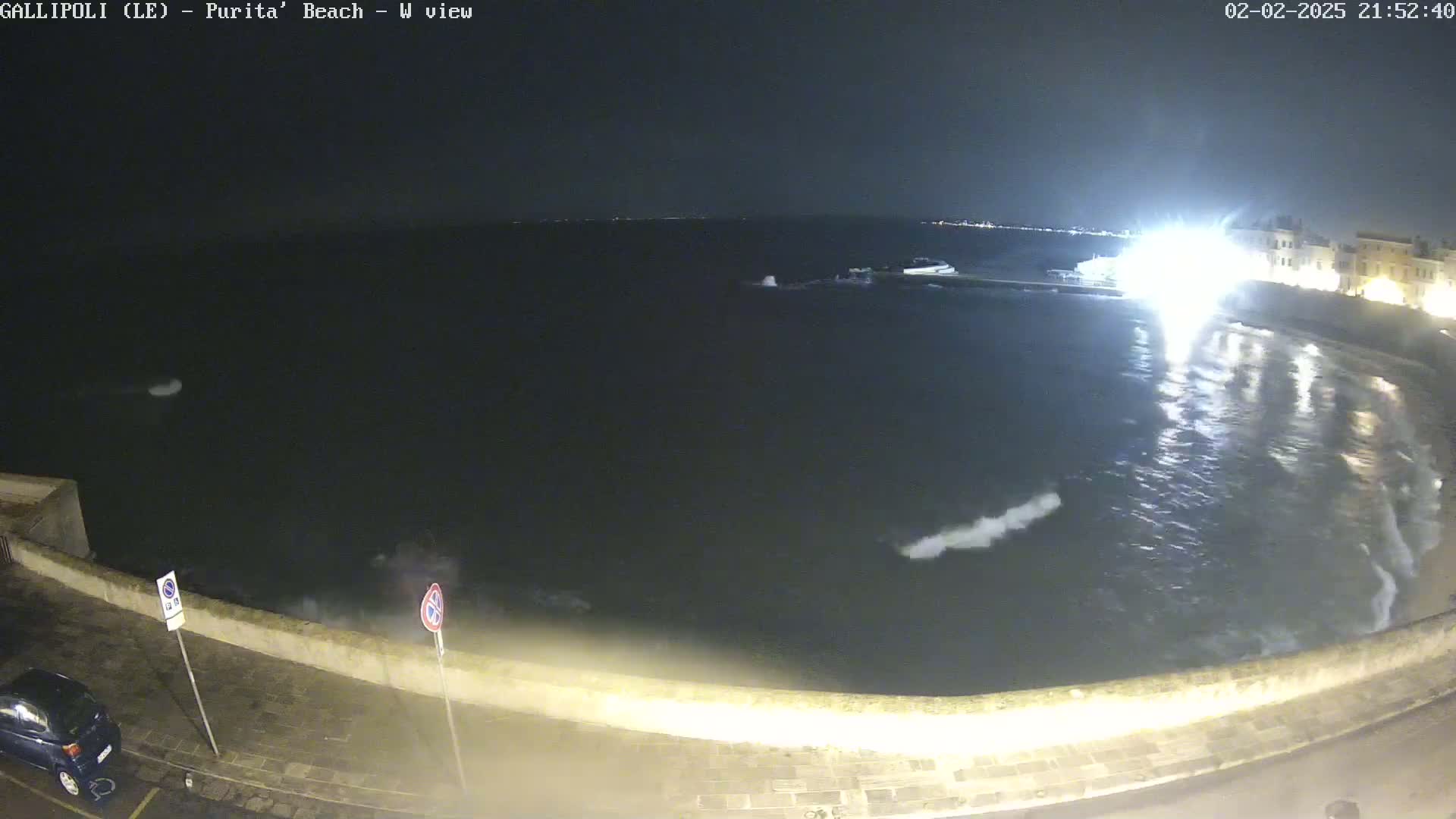 A nighttime view of a dark, wavy sea lapping against a low stone wall, with a few lights visible on the shore.
