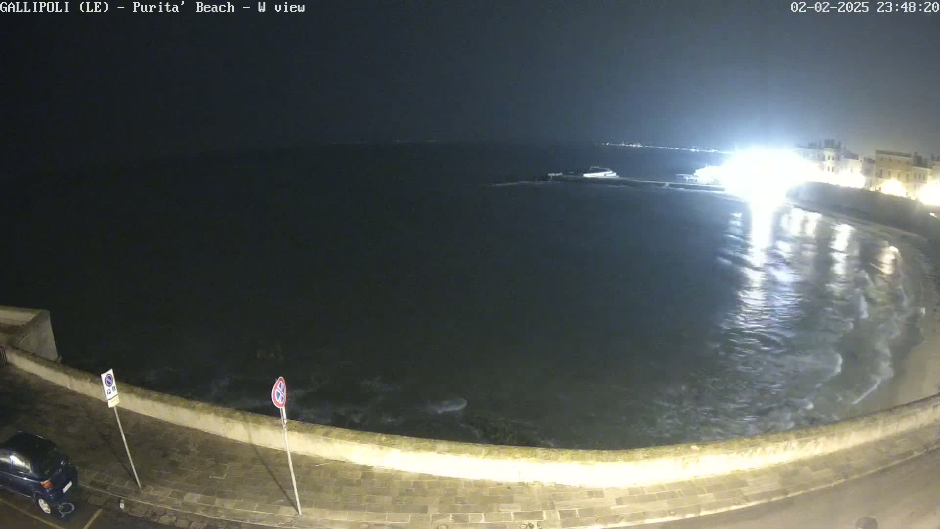 A nighttime view of a calm sea lapping a stone wall, with some buildings and lights visible in the distance.