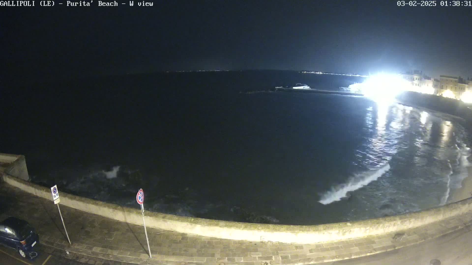 A nighttime view of a calm sea with small waves lapping against a stone wall, a parked car visible on the street above the wall, and buildings illuminated in the distance.