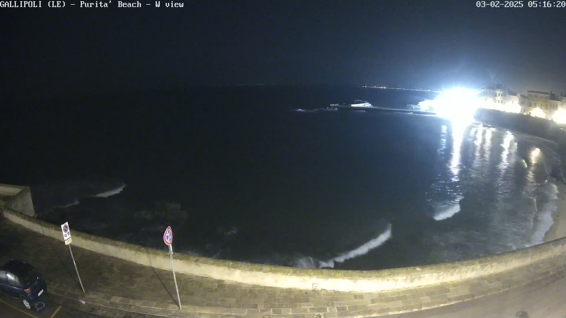 A nighttime, seaside view shows dark water with small waves lapping against a low stone wall, with lights visible across the water and along the shoreline.