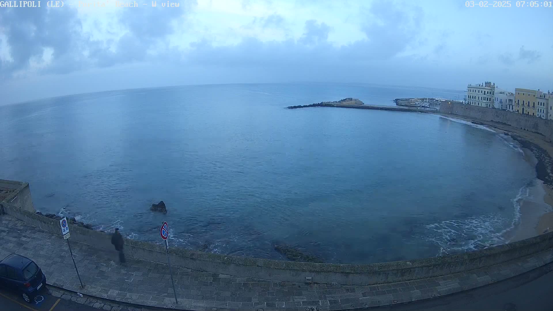 A partly cloudy morning shows a calm sea lapping a curved shoreline with buildings and a single person walking along a paved path.