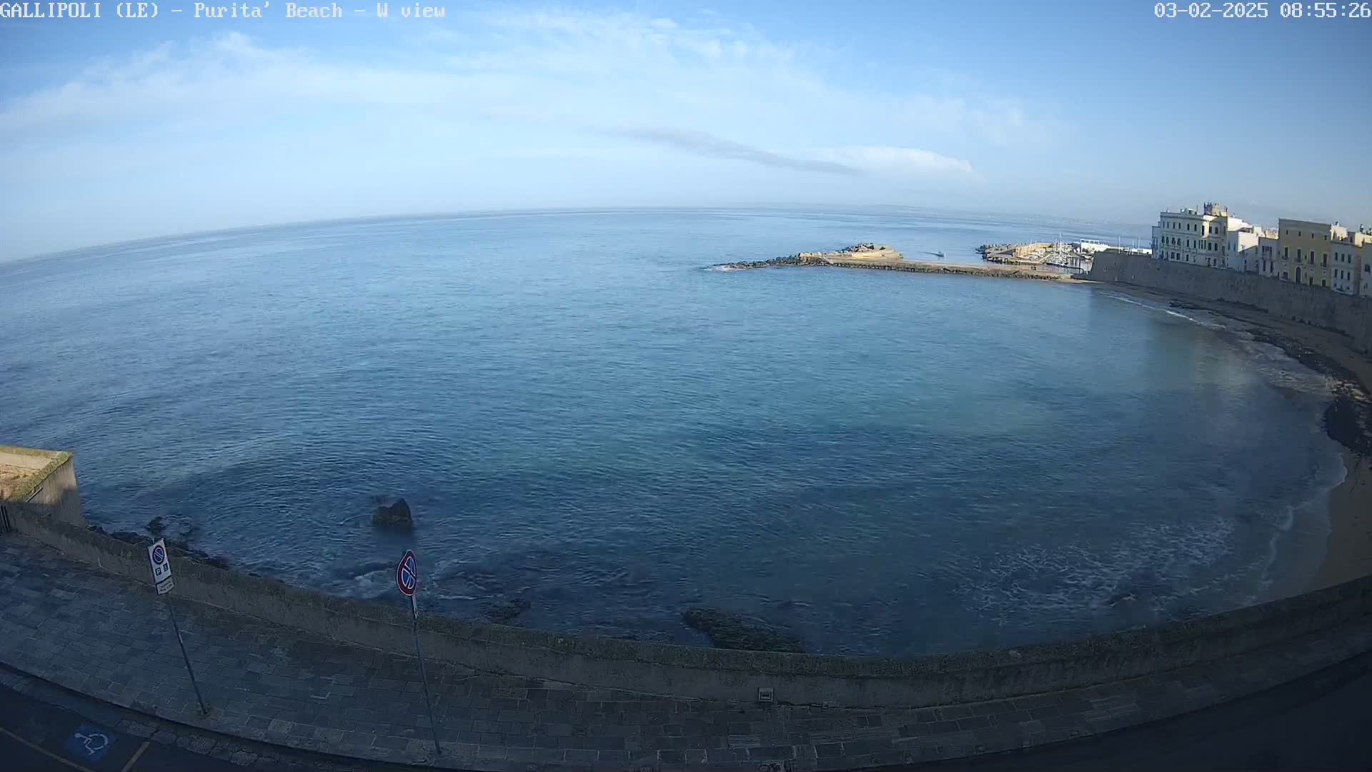 A coastal view shows a calm, blue sea lapping gently at a paved shore next to a low stone wall with buildings in the background under a mostly clear sky.